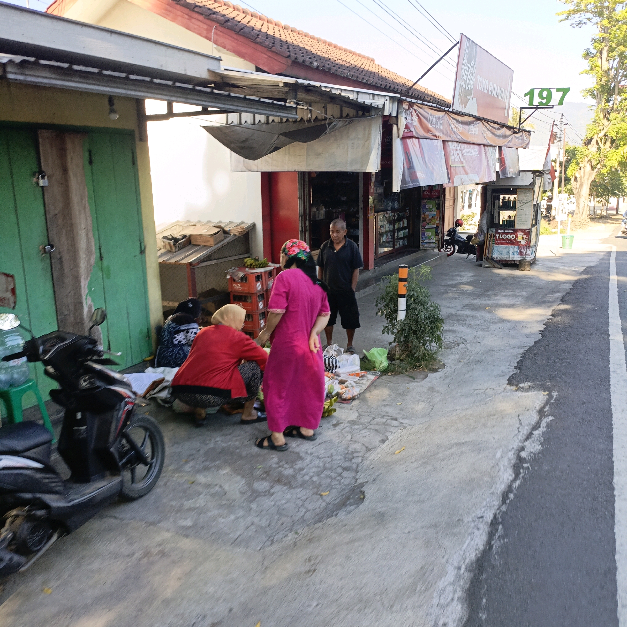 In the city of Gedangan, Indonesia, a street scene unfolds in front of a shop. A red-roofed building with an awning extends over the sidewalk. Three individuals are present. One man, wearing a dark polo shirt and shorts, stands near the shop entrance, looking to the side. Two women are crouched on the sidewalk near a red scooter parked on the left. One woman wears a bright red long-sleeved shirt and a headscarf, while the other is dressed in a vibrant pink dress and a patterned headscarf. Various items are spread out on the ground, including bags, what appear to be food items, and some crates. A tall green chair is visible to the left of the scooter, and a large water bottle sits on it. The pavement is cracked and weathered. A sign above the shop reads "TOKO GUNAWAN" and the number "197" is prominently displayed. The scene is bathed in sunlight, with shadows cast on the pavement.