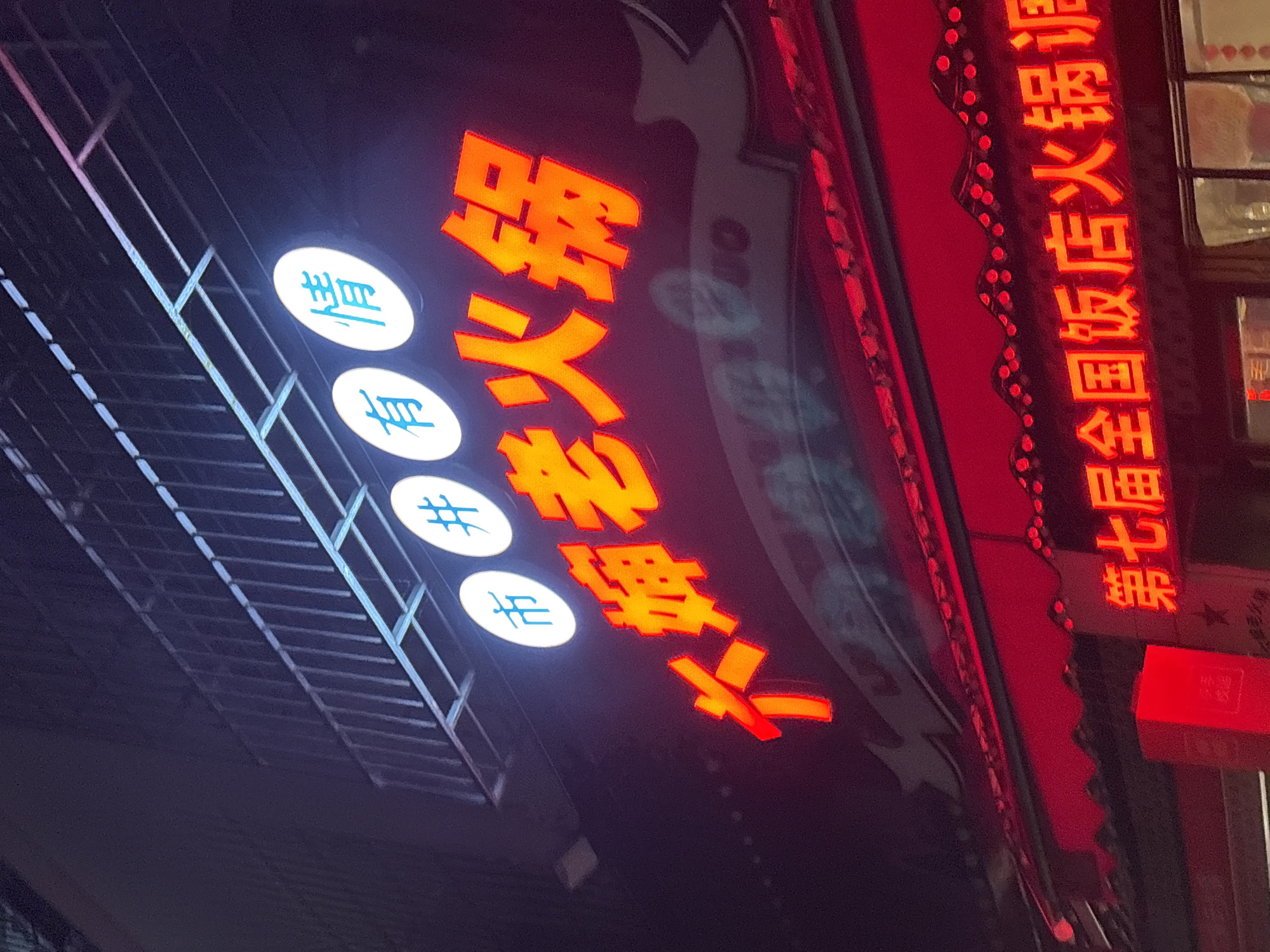 A nighttime photograph shows a brightly lit storefront in Nan'an District, Chongqing, China. The signage is in Chinese. In red neon on the right side, the characters say "Seventh National Hot Pot Food Festival". To the left, the orange-red characters are "Hot pot, many people, food". To the left again, the circular signs contain a symbol that represents "ear", two hashtag symbols, and a final unidentifiable symbol. The signage structure is made of metal. The red awning also contains decorative lights along its edges. A fire-extinguisher box is located on the right.