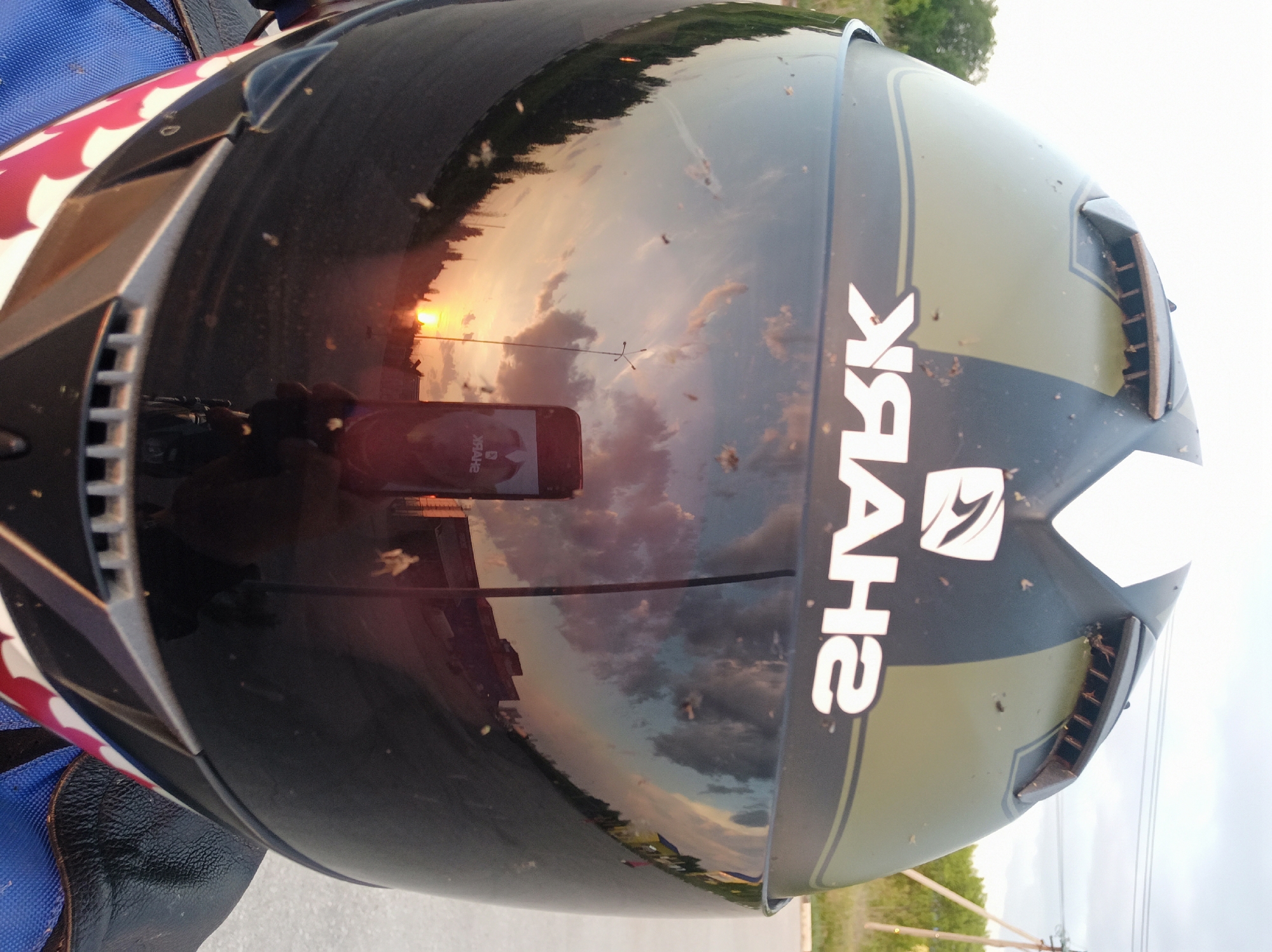 Here's a detailed description of the image:
**Scene:** An outdoor scene, likely a street or roadside, during sunset.
**Subject:** A motorcycle helmet is the main subject of the image. The helmet is black with an olive green accent stripe and the word "SHARK" in white. It has a white arrowhead design at the front. The helmet's visor is highly reflective, showing the reflection of the sunset, clouds, a telephone pole, and the photographer's cell phone. The helmet is dirty, with visible insect remains.
**Objects:**
* Motorcycle helmet
* Reflective visor
* Cell phone (reflected)
* Telephone pole (reflected)
* Street/road
* Trees in the background
**Environment:**
* Sunset, with the sun low on the horizon and clouds in the sky.
* The scene is likely near a road, as evidenced by the road reflection in the visor.
* The presence of a telephone pole further suggests a roadside location.
**Location:** While the image provides no explicit markers to positively identify the exact location as Perm, Russia, the landscape, time of day, and objects present in the reflection make the possibility plausible. The reflections show a typically Russian landscape, with electricity lines in view and what looks like typical architecture.