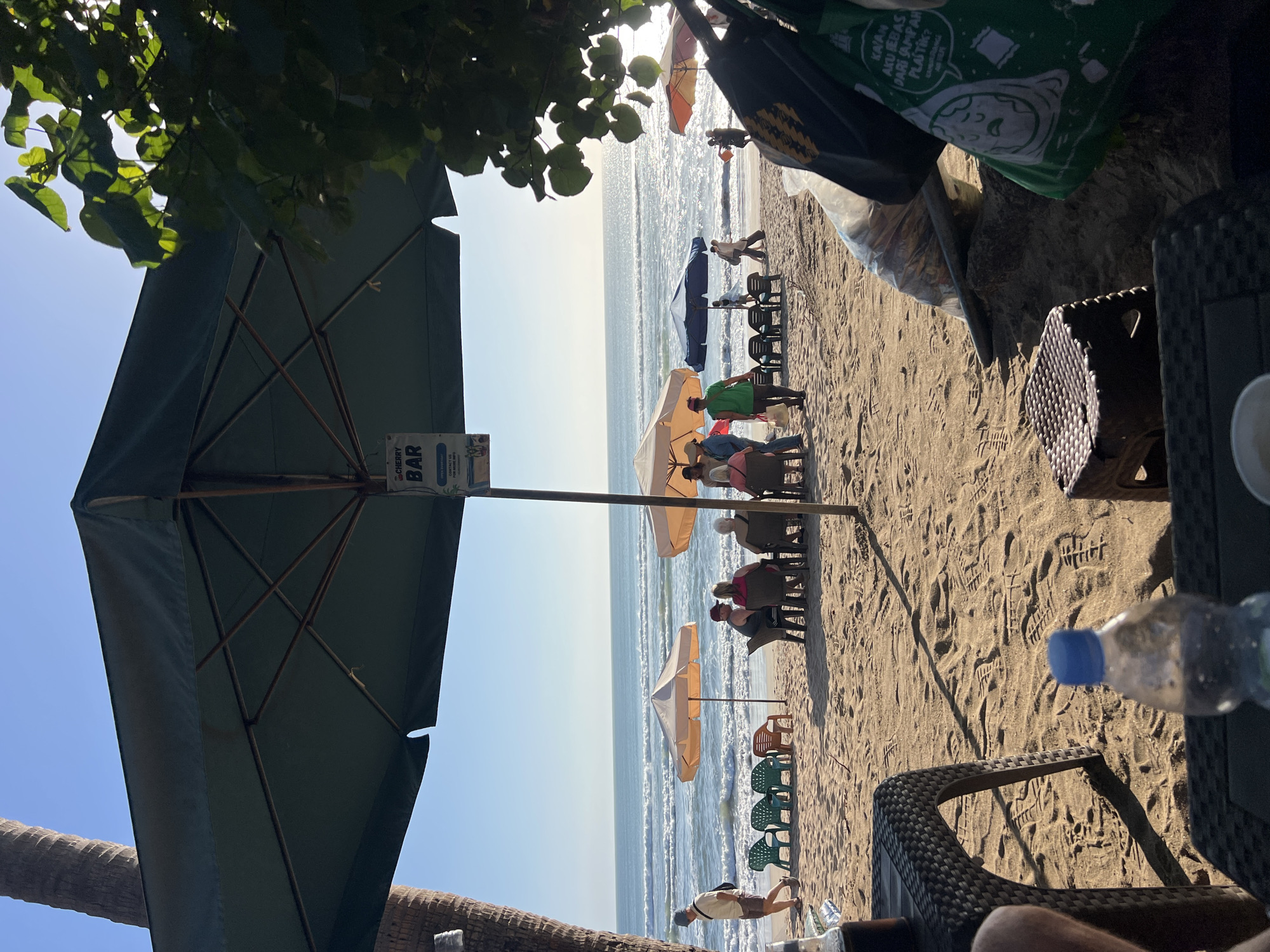 The image depicts a beach scene in Kuta, Indonesia, on a clear, sunny day. The foreground features two woven plastic tables and stools on the sand, with a bottle of water and a glass of dark liquid on one of the tables. Beyond the tables, a large green umbrella provides shade over several patrons seated at tables. Further back, beachgoers stroll along the shore, some are seated at various tables under umbrellas, and others are walking in the distance near the ocean. The ocean itself displays gentle waves and a shimmering surface due to the sunlight. Palm trees are visible on the left, and lush green foliage drapes over the right side of the frame, partially obscuring the view. A sign for "CHERRY BAR" is visible hanging from the umbrella pole. The sand is imprinted with numerous footprints.