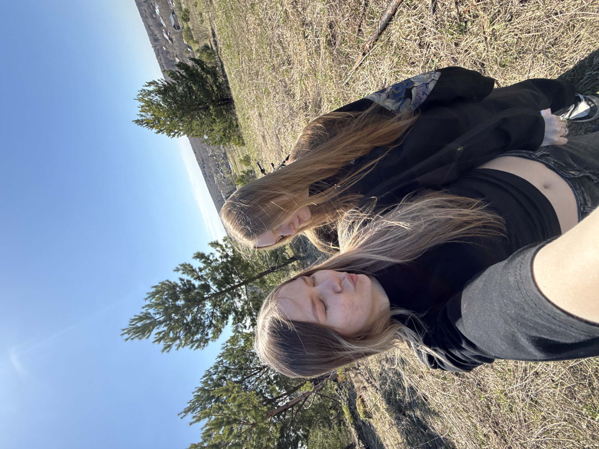 Two young women are lying on their backs in a grassy field. The bottom woman is wearing a black shirt and dark gray or black jeans. The top woman is wearing a black jacket with a floral pattern on the sleeves. The field is dry and brown, with some green trees and bushes in the background. The sky is blue. The photo appears to have been taken in Zhigalki, Russia.