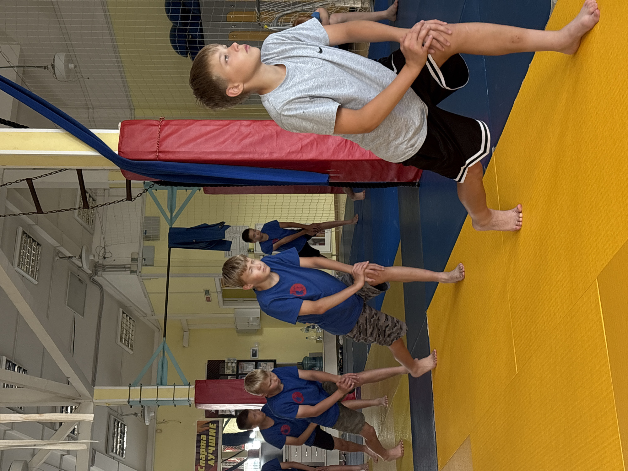 The image depicts an indoor gymnasium with a yellow and blue matting floor. In the foreground, a young Caucasian boy wearing a grey t-shirt and black shorts with white stripes is stretching his left leg forward, with his hands clasped on his bent left knee. His right leg is extended backward, and he is barefoot. Behind him, several other boys, also barefoot, are engaged in similar stretching exercises. They are wearing blue t-shirts and camouflage shorts. The gymnasium features a high ceiling with exposed beams and fluorescent lighting. Red and blue padded structures are visible, along with a net in the background. A banner with Russian text, "Спарта Лучшие" (Sparta Best), is displayed on the wall. This scene is likely taking place at a sports facility in Chaikovsky, Russia, given the context.