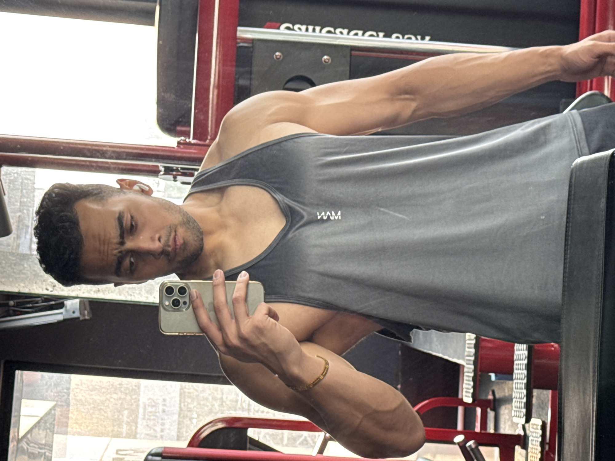 A man is posing for a selfie in what appears to be a gym. He is wearing a gray tank top and holding an iPhone. His muscular arms are prominent. The gym equipment behind him is predominantly red and black. The background visible through a window suggests a cityscape, possibly in Kolkata, India, given the image's origin.