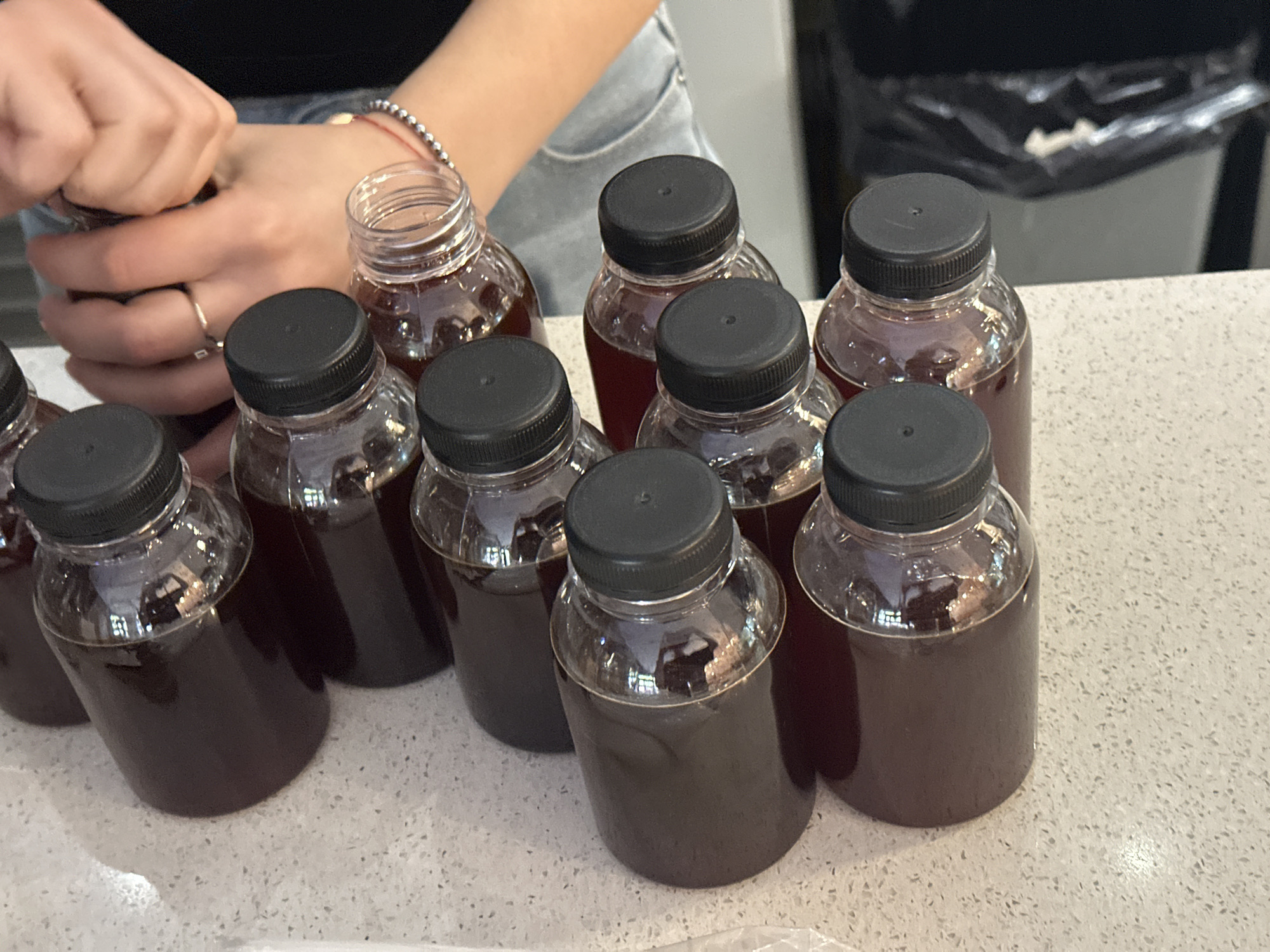 On a light-colored, speckled countertop, there are nine small, clear plastic bottles filled with a dark liquid, likely coffee or tea. Each bottle has a black screw-on lid. A person, seen from the torso up, is closing the lid on the topmost bottle. This person is wearing a black top and light-wash jeans, and they have a silver bracelet on their wrist and a ring on their finger. In the background is a black trash bin with a clear plastic bag. It is not possible to ascertain the location as Novi Sad, Serbia from the image.