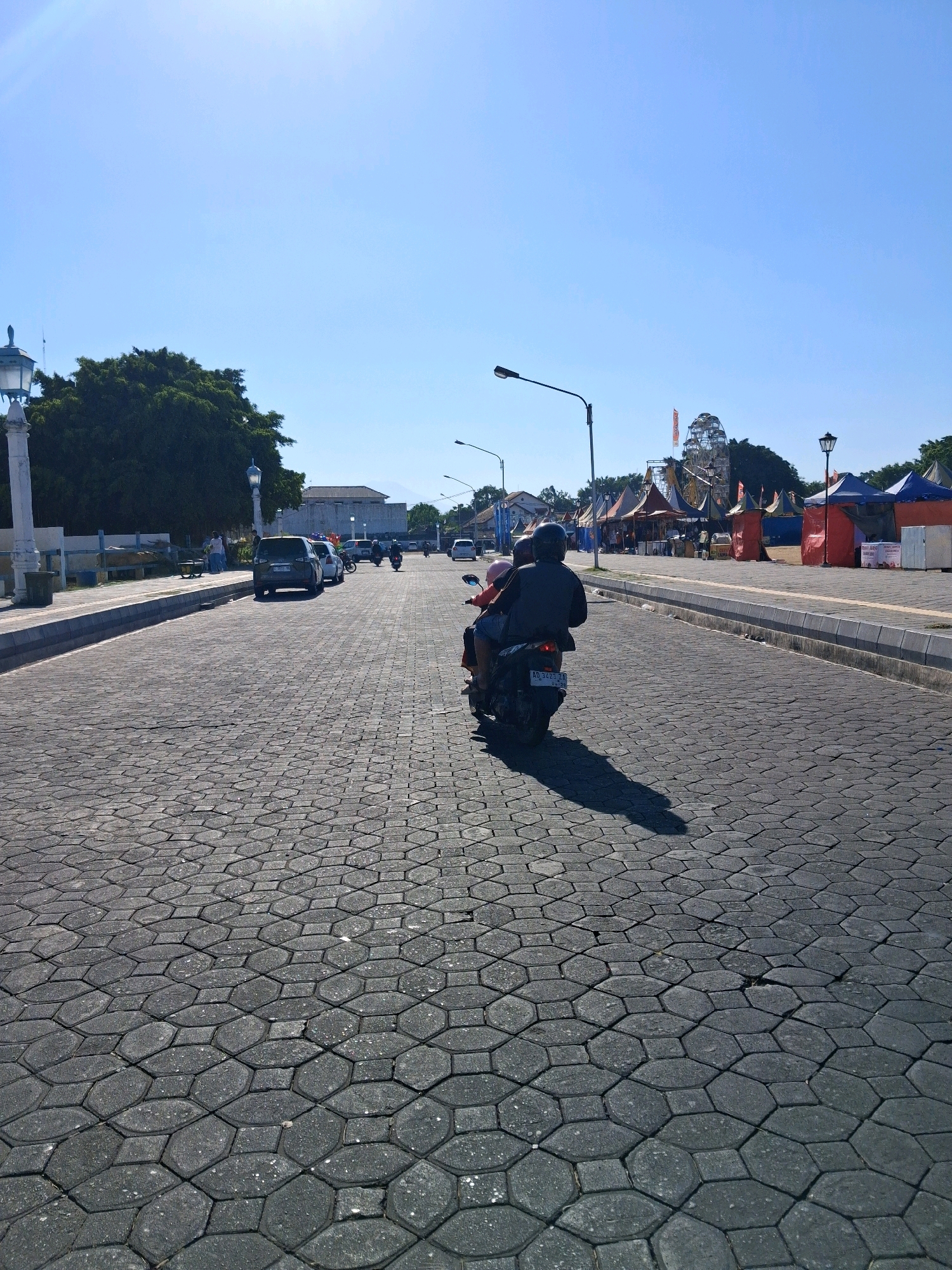 A perspective shot from behind two people on a motorcycle on a cobblestone street with a clear blue sky. The motorcycle is traveling away from the viewer towards a row of tents and a Ferris wheel. The street is paved with hexagonal cobblestones and is lined with parked cars and streetlights. In the distance, there are buildings and trees. The Ferris wheel and tents suggest a fair or market setting.