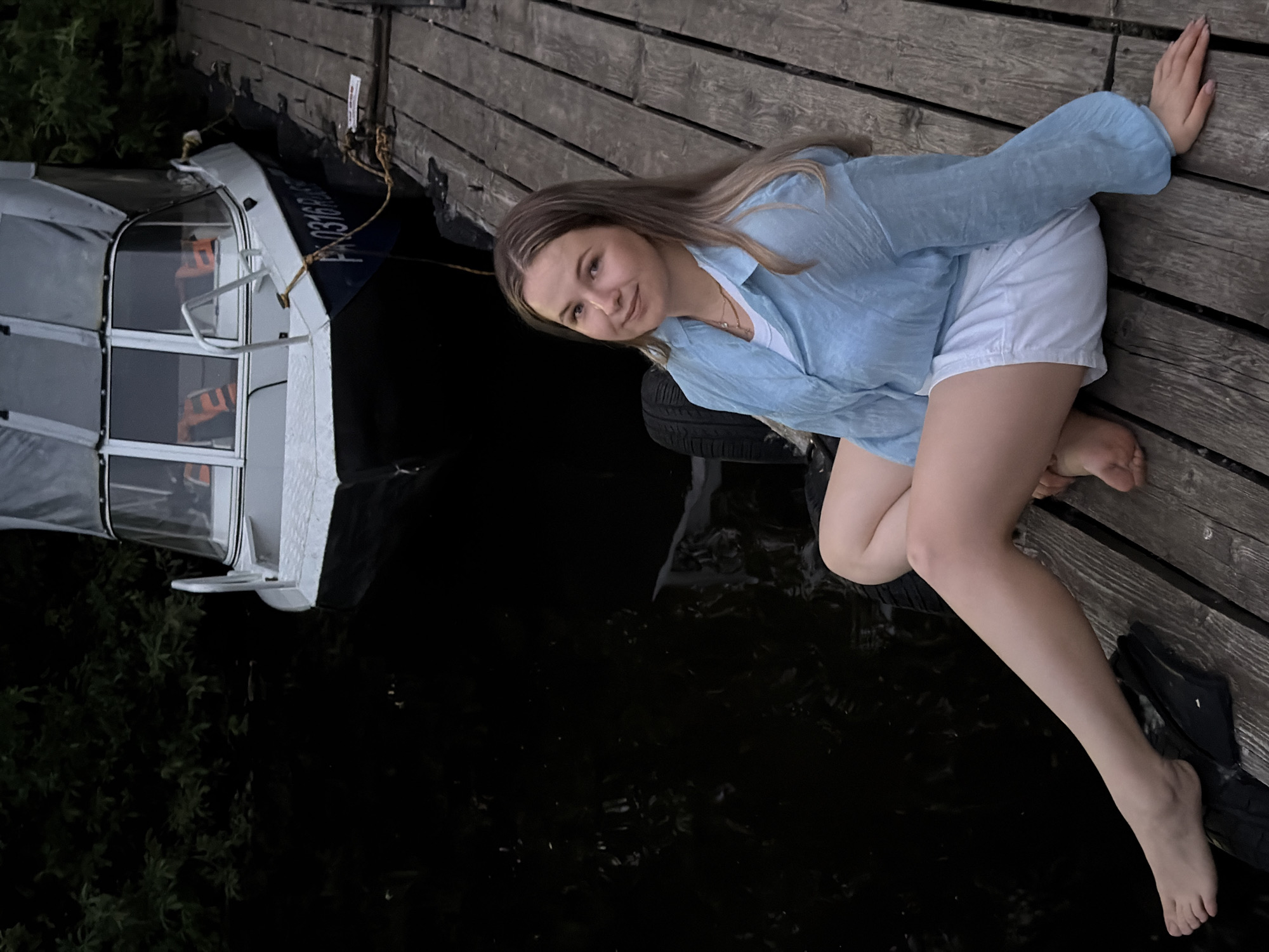 In Chaikovsky, Russia, a young woman with blonde hair sits on a wooden pier next to a dark body of water. She is wearing a light blue translucent shirt and white shorts. Her legs are crossed, and her feet are bare. To her right, a boat is docked. The boat is white with a grey cover and has orange life vests visible inside. The pier is made of weathered wooden planks. Trees are visible in the background.