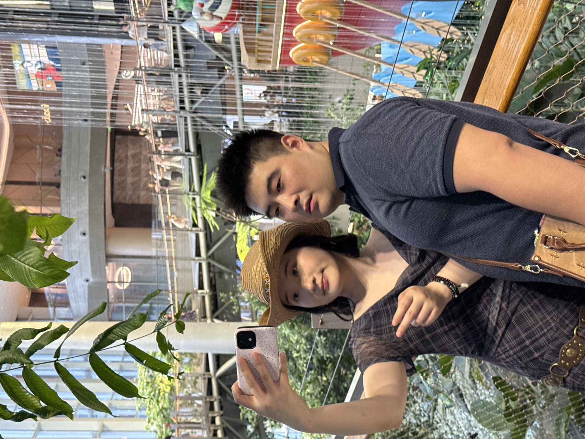 In an indoor atrium, possibly a shopping mall or entertainment venue in Liangjiang, China, a young Asian woman and a young Asian man are posing for a selfie. The woman, wearing a straw cowboy hat and a plaid dress, holds up a smartphone with her left hand. She points her right index finger towards the man. The man, wearing a navy blue polo shirt, stands beside her. In the background, there are multiple levels of walkways and railings, with lush green plants and a large, decorative waterfall feature. People can be seen on the walkways in the distance. A large, colorful boat-like structure with decorative rings is visible on the right side of the frame.