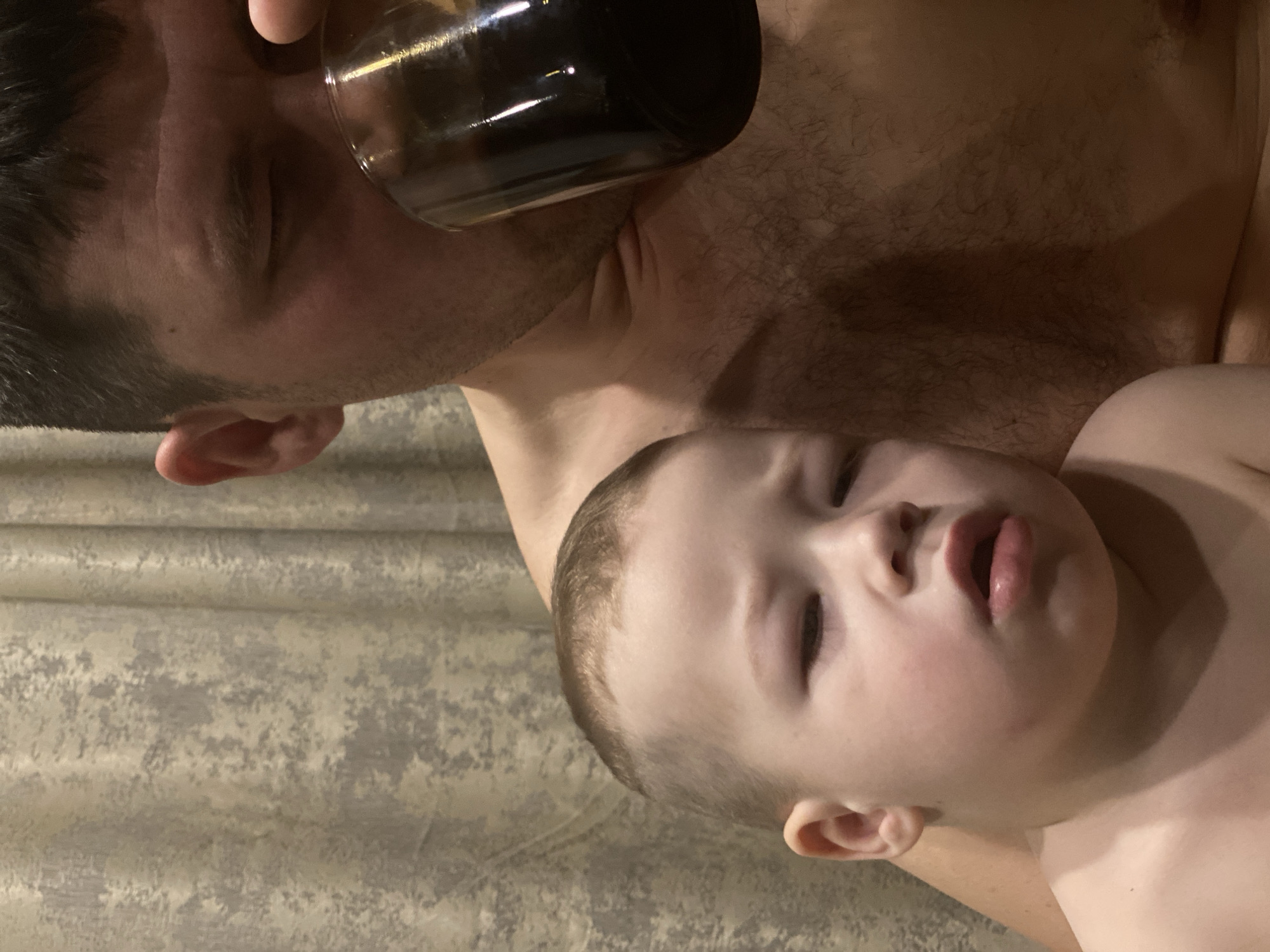 A close-up, intimate shot shows a man drinking from a dark glass container while a young child rests on his chest. The man has dark hair, fair skin, and closed eyes as he brings the glass to his mouth. The child, also fair-skinned with light hair, appears drowsy or asleep with slightly parted lips. The man's chest is visible, showing some hair. The background is a blurred, patterned wall. It is not possible to determine the location of the image as Sochi, Russia from only the given information.