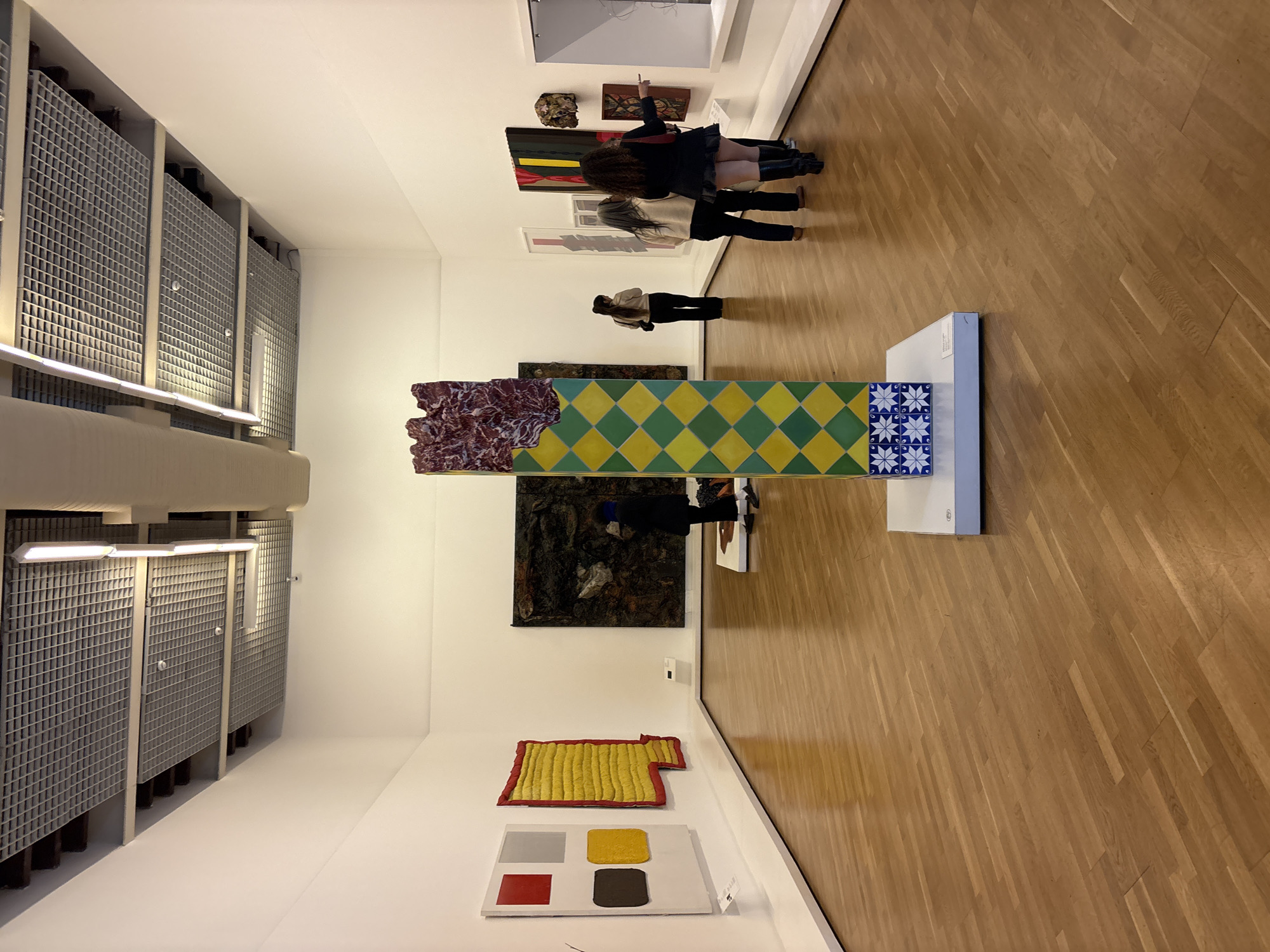 The image shows an art gallery. The gallery walls are white and adorned with various contemporary artworks. Several visitors are viewing the art, including a group of two women. One of the pieces is a sculpture with a marbled rock-like element and a yellow and green diamond-patterned section. Another piece is a large abstract painting with dark colors. There's a yellow textile artwork and a painting with red, grey, yellow and brown squares. The floor is made of wood. The gallery has a modern ventilation system visible on the left side. The location is São Paulo, Brazil.