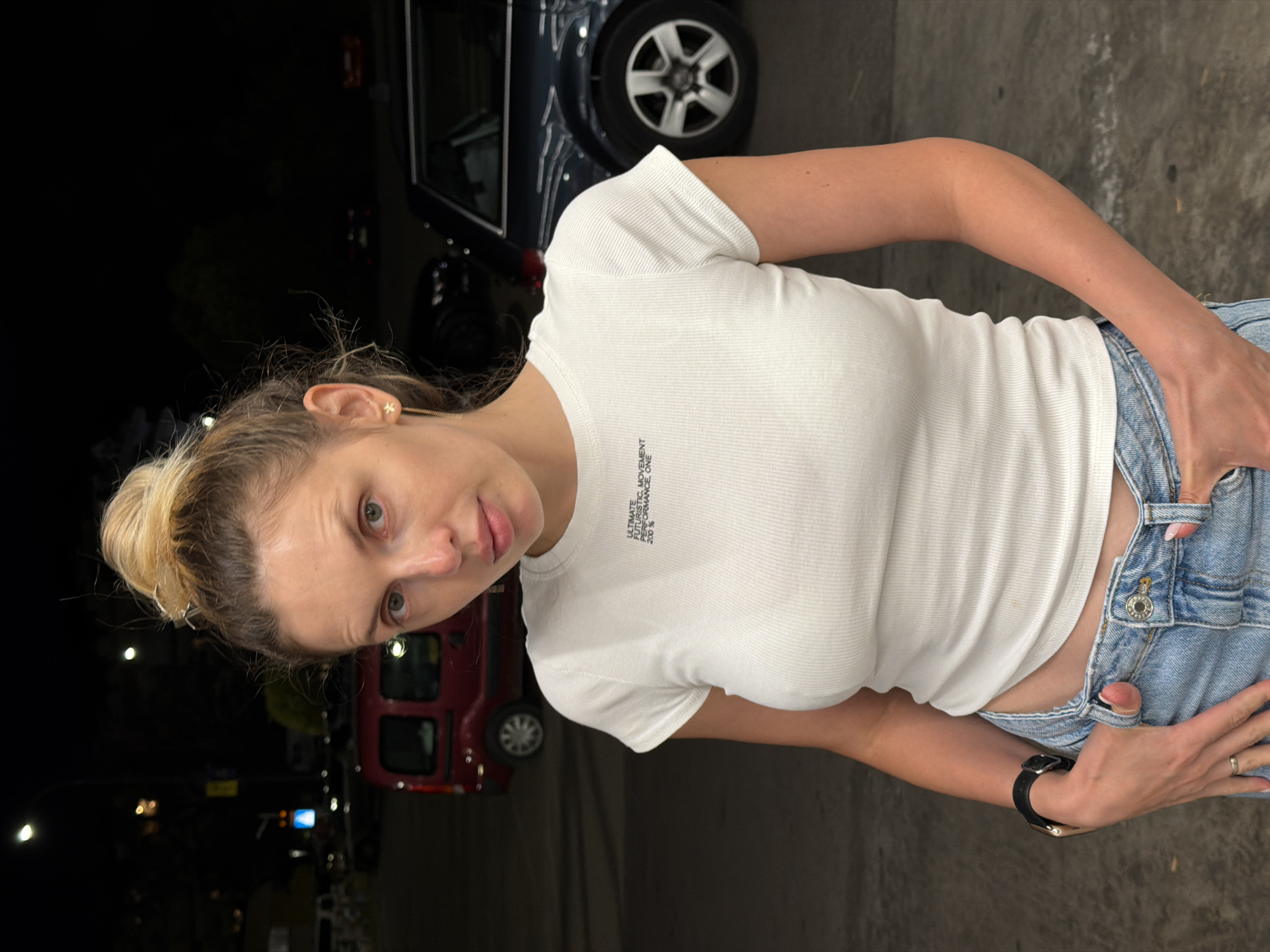 A blonde woman with her hair in a bun stands outdoors at night, looking at the camera. She is wearing a white ribbed t-shirt and light blue jeans. Her left hand is in her jeans pocket, and her right hand is also in her pocket. A black watch is on her left wrist. Behind her, a red van and a dark-colored SUV are parked. Streetlights illuminate the scene. The location is the parking lot of the "Maxi" supermarket on the border of Budva, Montenegro.