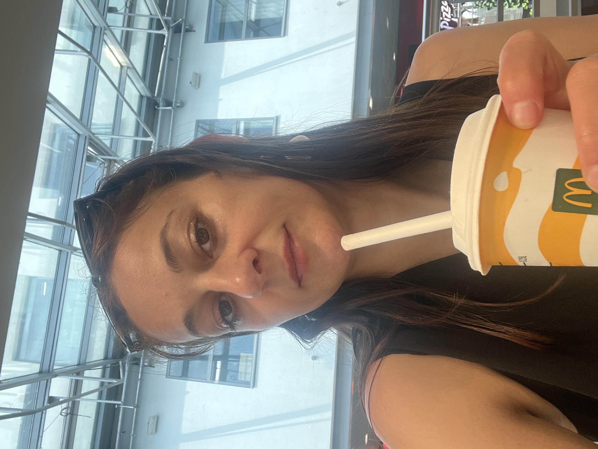 This is a close-up, selfie-style photograph of a young woman holding a McDonald's drink cup. The image is oriented vertically. The woman is looking directly at the camera with a neutral expression. She has long, dark brown hair that falls around her face and shoulders. She is wearing a black top. A white straw is visible in the cup, which has the McDonald's logo and branding on it, featuring yellow and white colors.
In the background, a large glass-paned ceiling with a metal frame is visible, suggesting an indoor location with a lot of natural light. The structure appears to be part of a modern building, possibly a shopping mall or a public transport hub. The light streaming through the ceiling creates bright highlights and shadows on the woman's face and in the background. Some windows and a white wall are also visible in the background, with hints of greenery and signs, suggesting an urban setting. The overall lighting indicates it is daytime. There is text visible on the cup.