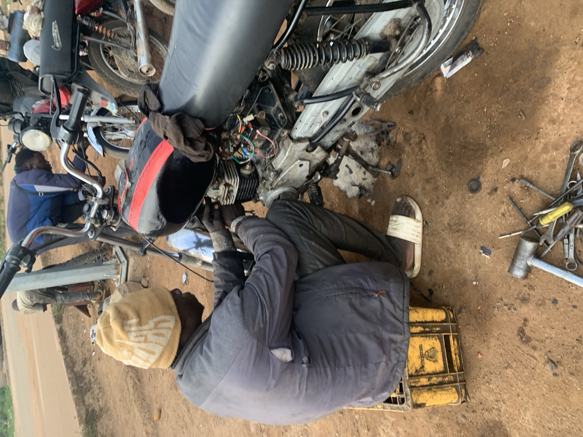 A man is working on a motorcycle in what appears to be a repair area in Barakin Lerawi, Nigeria. The man, wearing a beige beanie and a grey jacket, is seated on a yellow plastic crate and is leaning over the engine of a black motorcycle. Wires and mechanical components are exposed on the motorcycle's engine. In the background, another man in a light-colored shirt is bent over a different motorcycle. The ground is a reddish-brown dirt, with scattered tools including wrenches and a hammer visible in the foreground. The overall environment suggests an informal outdoor workshop.