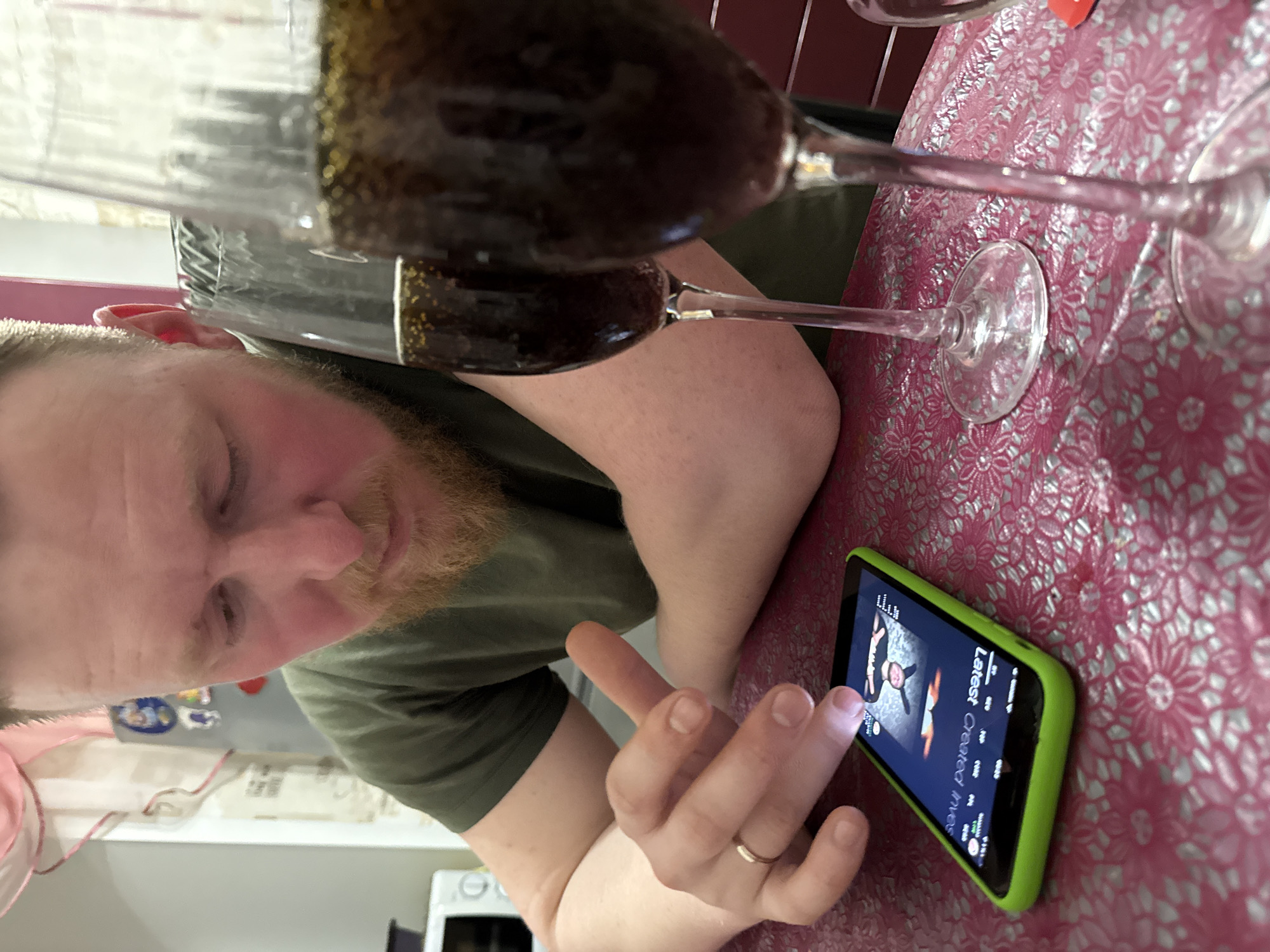 A fair-skinned man with a beard is looking down at a smartphone with a green case. He is touching the screen with his index finger. He is wearing a dark green t-shirt and a gold wedding band on his left ring finger. Two stemmed glasses filled with a dark liquid are placed on a red and silver floral patterned surface. The background is blurred but appears to be a kitchen setting with white cabinets and appliances. The location is likely within the city of Perm, Russia.
