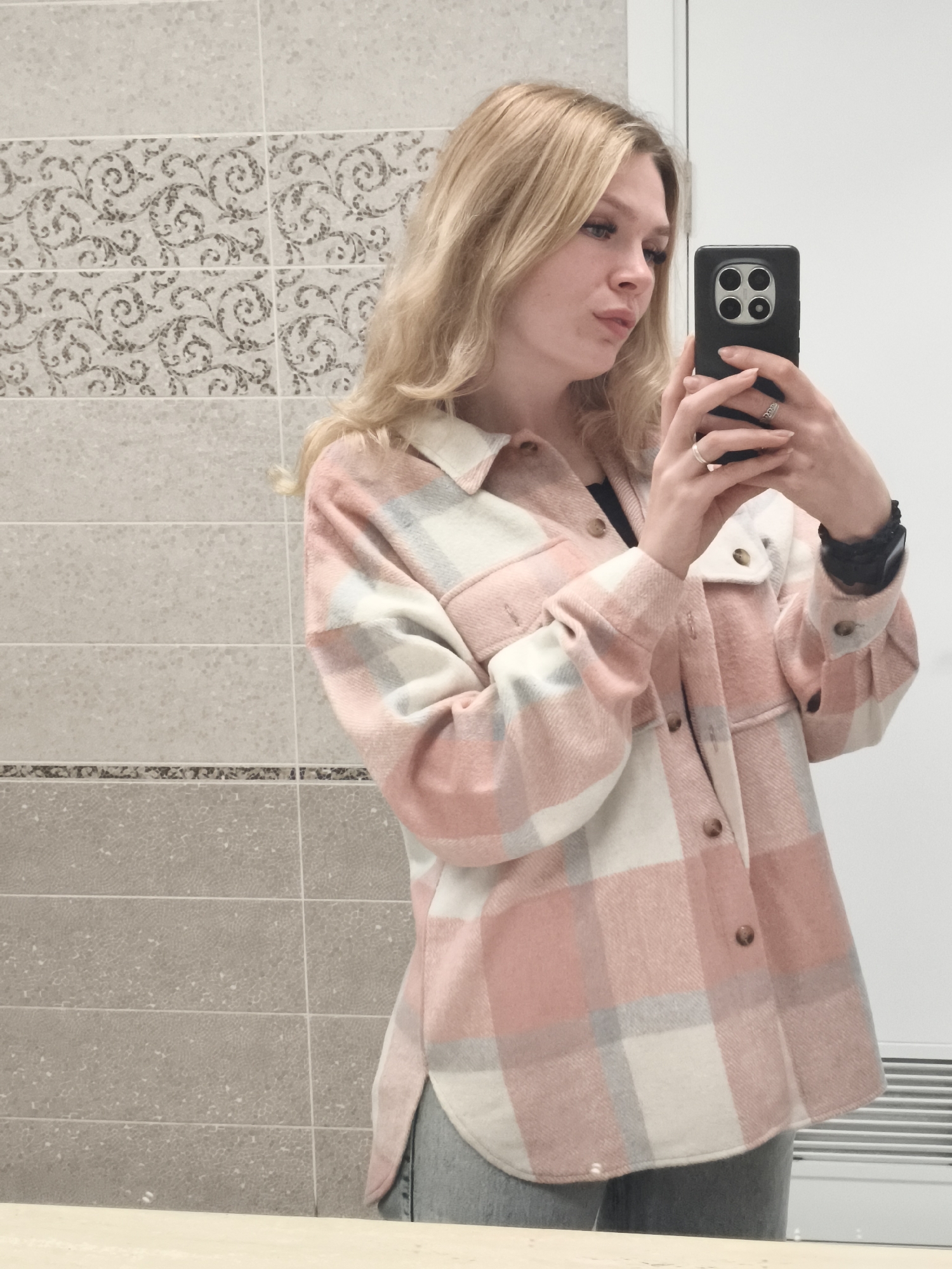 A fair-skinned woman with blonde wavy hair is taking a selfie in a restroom. She is wearing a pink and white plaid shirt-jacket over a black top and gray pants. She is holding a black smartphone with four circular camera lenses in her right hand. She is wearing two rings, one on each hand, and a black wristband on her left wrist.
The background consists of a tiled wall with two different types of tiles: a gray tiled wall with light-colored patterned tiles. Part of a white door and heater are visible to the right.
There is no evidence in the image to determine if the location is in the city of Izhevsk, Russia.