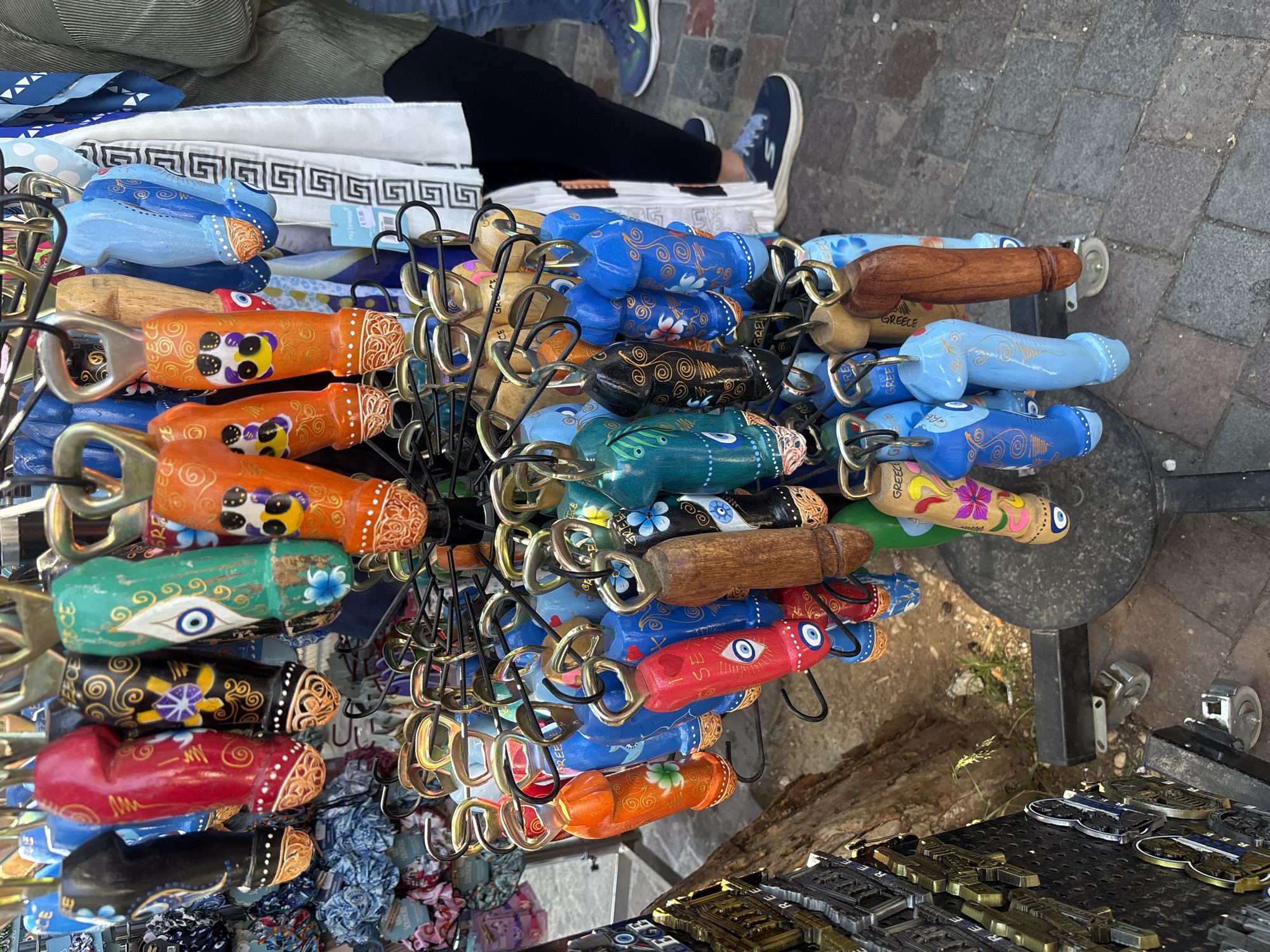 The image depicts an outdoor souvenir stall, likely situated in a pedestrian market area such as Plaka or Monastiraki in Athens, Greece. The primary subject is a black metal rotating display rack laden with numerous phallic-shaped wooden bottle openers or keychains. These items are brightly colored in red, black, green, orange, blue, and natural wood tones, featuring painted decorations like swirls, flowers, and the prominent Greek "evil eye" symbol; some are explicitly marked with "GREECE." To the left, a secondary display rack holds metallic rectangular souvenirs. Behind the main display, various colorful fabric items are visible. On the right side, a white scarf with a repeating black geometric pattern hangs from an adjacent rack, with a "my breeze" price tag indicating "15.00." Two individuals are partially visible on the right, standing on the paved ground: one wearing a light green long-sleeved shirt and dark pants, and another in blue jeans and blue sneakers, one of which has a distinct neon yellow swoosh. The ground is composed of gray paving stones, with sparse vegetation visible near the base of the display stands. The scene is illuminated by natural daylight.