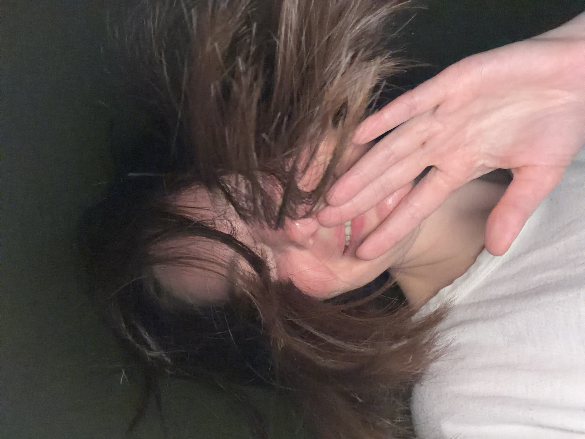A close-up shot shows a person lying down, face partially covered by brown hair and a hand. The person's skin is fair, and their lips are slightly parted, revealing teeth. They are wearing a white top. The background is a dark, indistinct green. No city or location can be determined from the image.