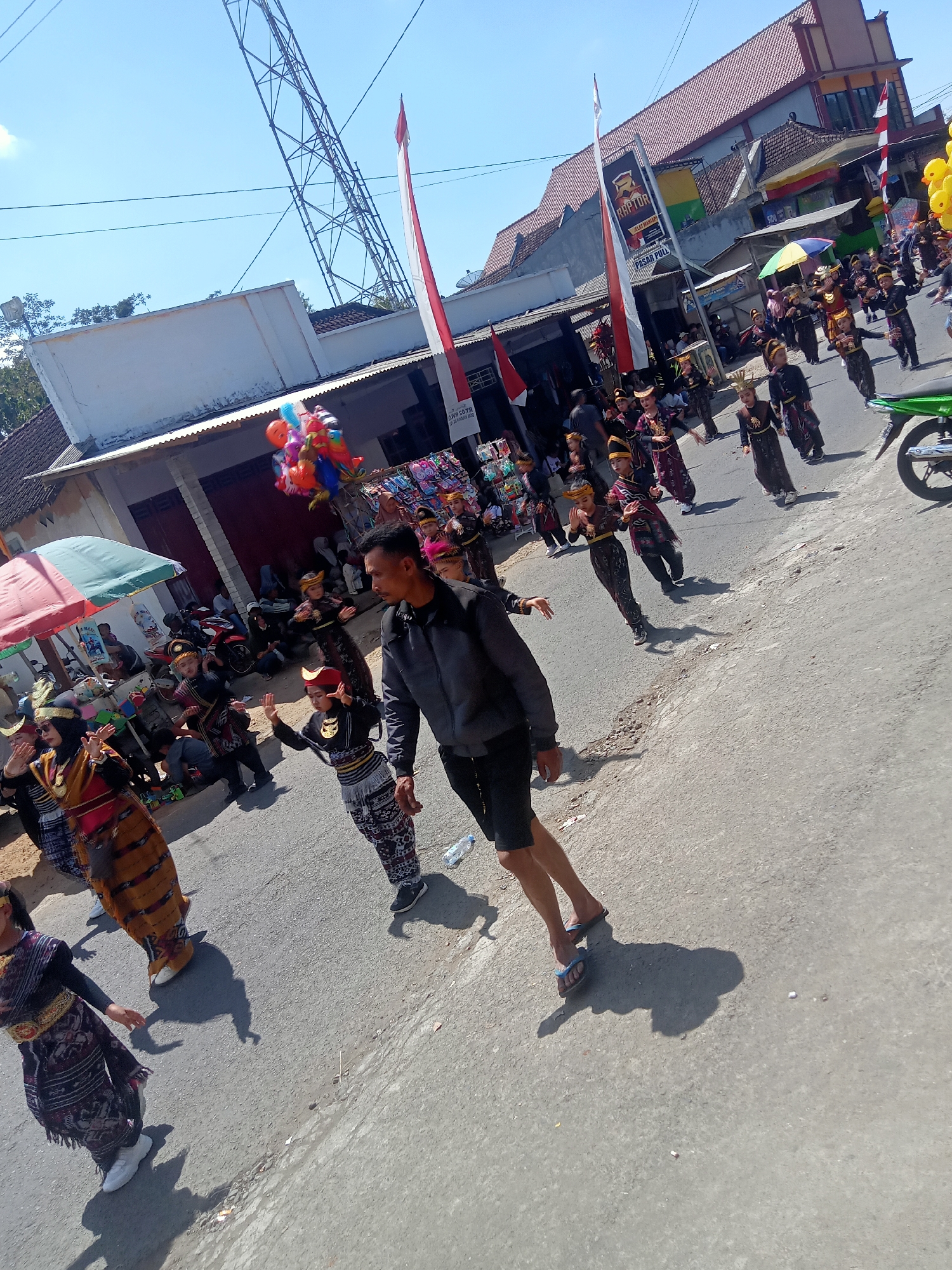 The image captures a street scene in Pule, Indonesia, likely during a cultural parade or event. A group of children, dressed in traditional attire, are the central focus of the scene, walking down a paved road. The children are adorned in dark uniforms with intricate patterns and headwear, some carrying decorative elements. Several adults are also present, some participating in the procession and others observing from the sides.
In the foreground, an adult male in dark athletic wear and flip-flops is walking, casting a prominent shadow. To the left, another adult woman in traditional clothing and a headscarf is also part of the parade. The street is lined with buildings, some appearing to be shops with awnings and open doorways. A tall communication tower is visible in the background, and national flags of Indonesia (red and white) are displayed, some on poles and others attached to buildings. There are also balloons, some held by an unseen person, adding to the festive atmosphere. Motorcycles and other parked vehicles are scattered along the roadside. The sky is clear and blue, indicating a sunny day. The overall impression is one of a vibrant community celebration.