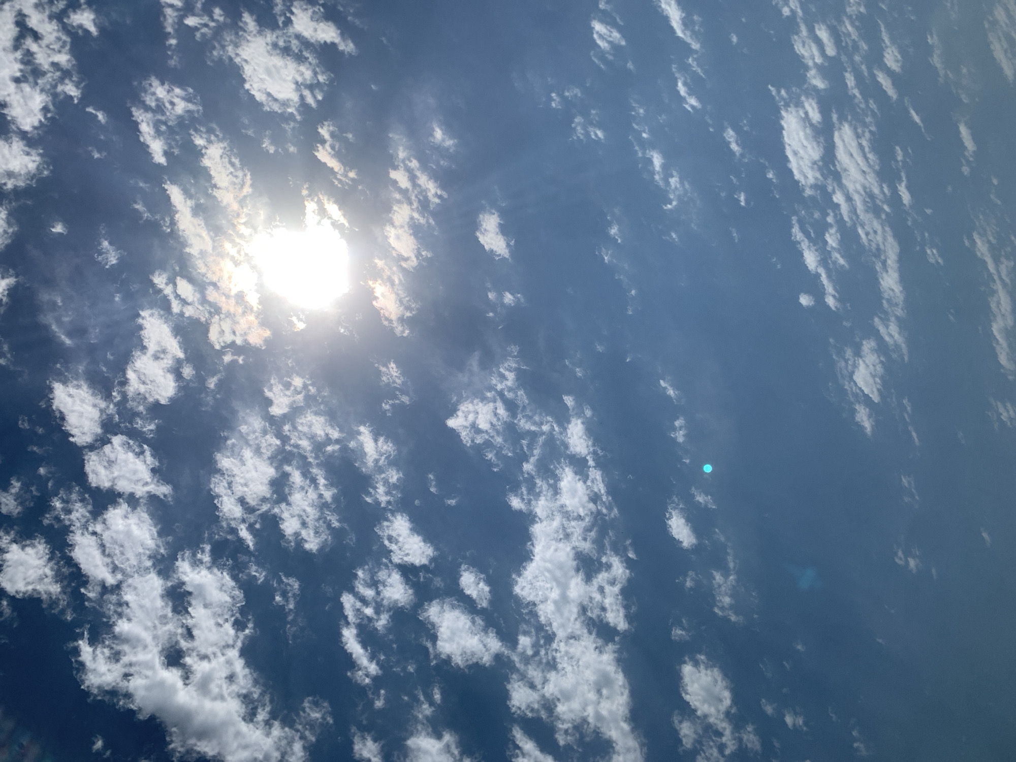 A bright sun shines through scattered white clouds against a clear blue sky. The sun is positioned in the upper center of the frame, casting rays of light that extend downwards and outwards. The clouds are varied in size and density, with some appearing as wispy streaks and others as more defined puffy masses. The overall impression is of a bright, sunny day. The image location is specified as Khazar Raion, Azerbaijan. There are no people, subjects, or objects in the scene other than the sky and clouds. There are no actions or interactions depicted. The environment is exclusively the sky.