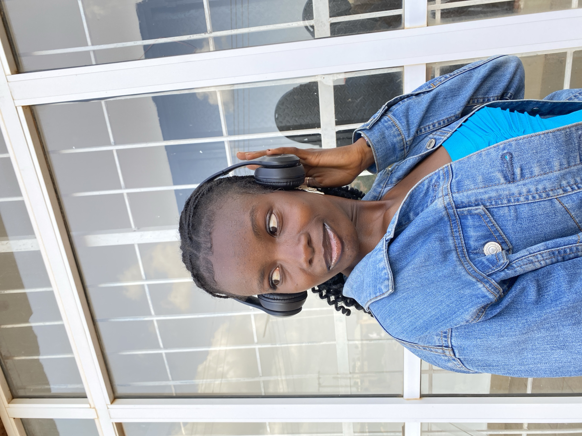 A medium shot shows a young Black woman wearing a denim jacket and a bright blue top. She has braided hair and is wearing large black headphones, with her right hand touching them. The woman is looking off to her left. She is standing in front of a large window with a grid pattern, and there are reflections of clouds in the glass. The background is out of focus. This image was taken in Bar Jirgi Summa, Nigeria.