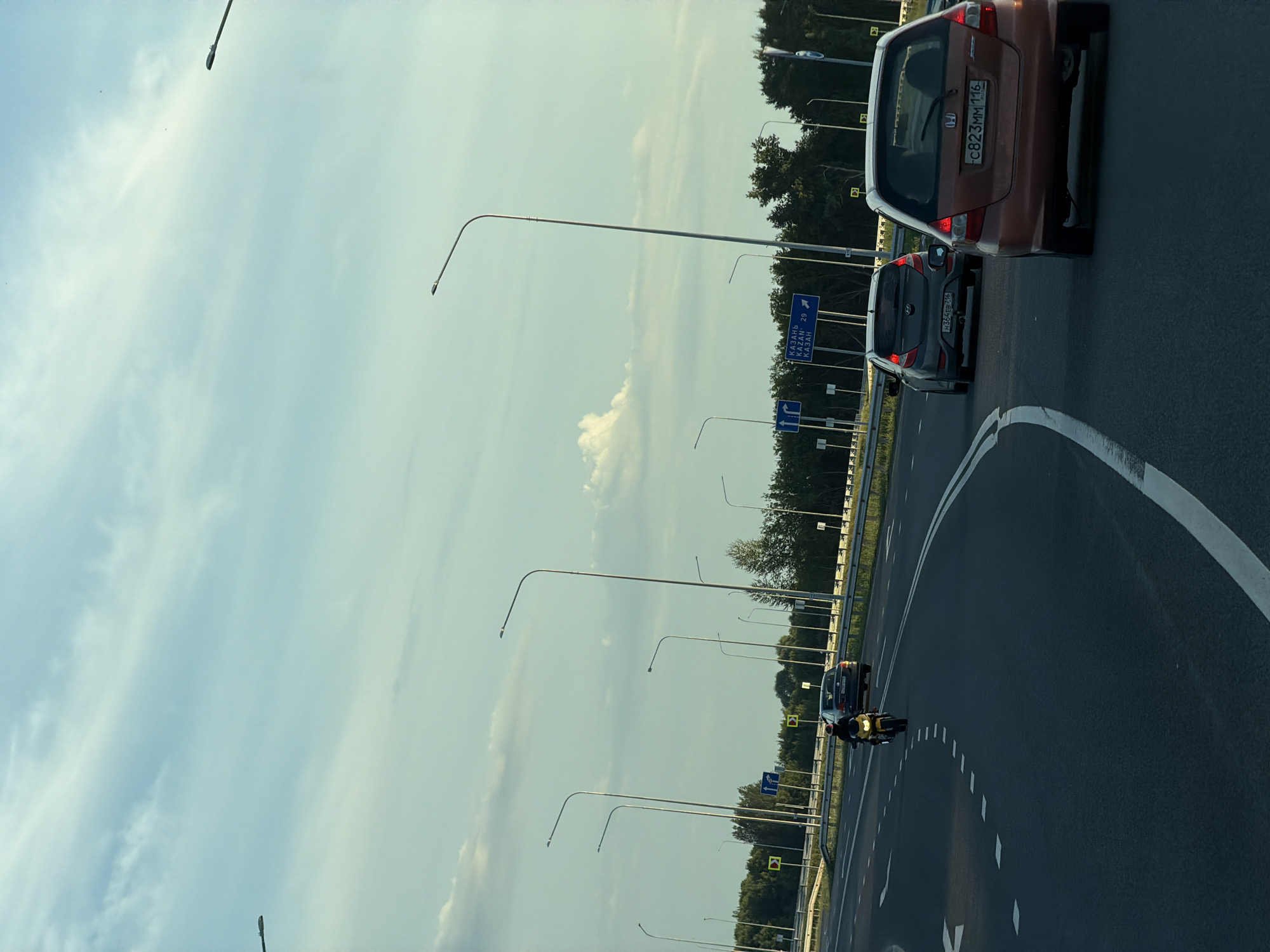 The image shows a highway scene with several cars and a motorcycle. In the foreground, an orange Honda Fit is driving on the right lane. A dark gray Hyundai Solaris is in the left lane, ahead of the Honda. In the distance, a motorcycle with a rider is on the left lane. The road is a multi-lane highway with streetlights lining the sides. The sky is partly cloudy with a light blue hue. There are trees and foliage visible beyond the highway barrier. A road sign in the distance indicates directions to "Kazan" with distances. The license plate of the orange Honda is "C823MM 116". The license plate of the Hyundai is "H364EB 716". The location is identified as Obukhovo, Russia.