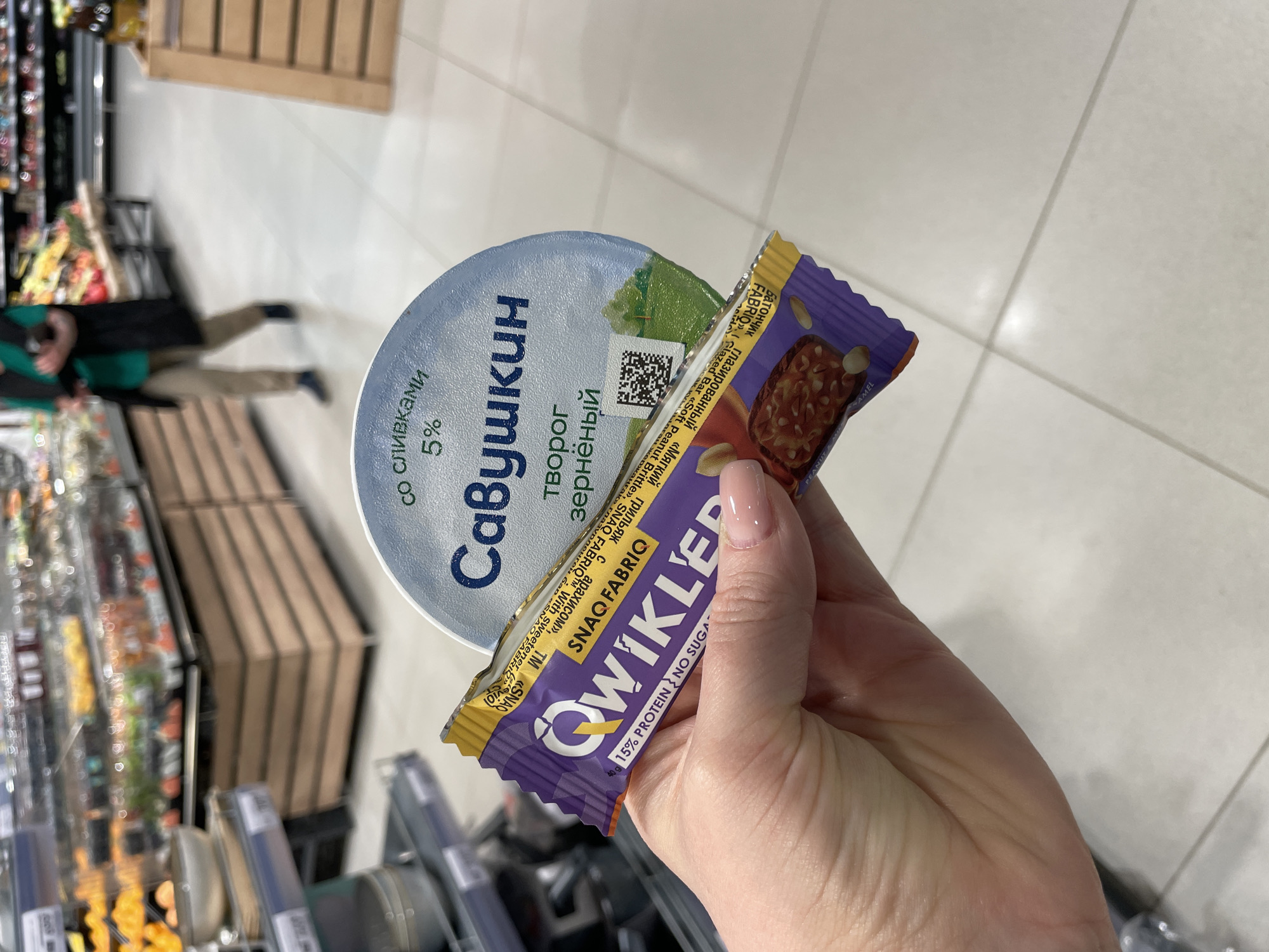 A close-up, slightly rotated shot depicts a person's hand holding two food items inside a brightly lit supermarket in Saint Petersburg, Russia. The hand, featuring polished nails, holds a round plastic container of cottage cheese and a packaged snack bar.
The container is a light blue lidded cup of "Савушкин" (Savushkin) brand cottage cheese, specifically "творог зернёный со сливками 5%" (cottage cheese with cream 5%). A QR code is visible on its green label.
Held next to it is a purple and yellow packaged snack bar from "SNAQ FABRIQ QWIKLER". Text on the wrapper indicates it's a "Батончик глазированный «Мягкий грильяж с арахисом», ТМ «SNAQ FABRIQ»" (Glazed bar "Soft peanut brittle with peanuts", TM "SNAQ FABRIQ"). English text also states "Glazed Bar «Soft Peanut Brittle», SNAQ FABRIQ™ With Sweetener" and "40g 15% PROTEIN NO SUGA" (implying no sugar). The bar itself, partially visible, appears to be made with peanuts and caramel, consistent with the "PEANUT & CARAMEL" text on the packaging.
The background, blurred, reveals typical supermarket elements: light-colored tiled flooring, various shelves stocked with products, and wooden crates. A partially visible and blurred person in a green top and beige pants can be seen in the mid-ground, suggesting other individuals are present in the store. The scene captures an everyday moment of grocery shopping within a modern retail setting.