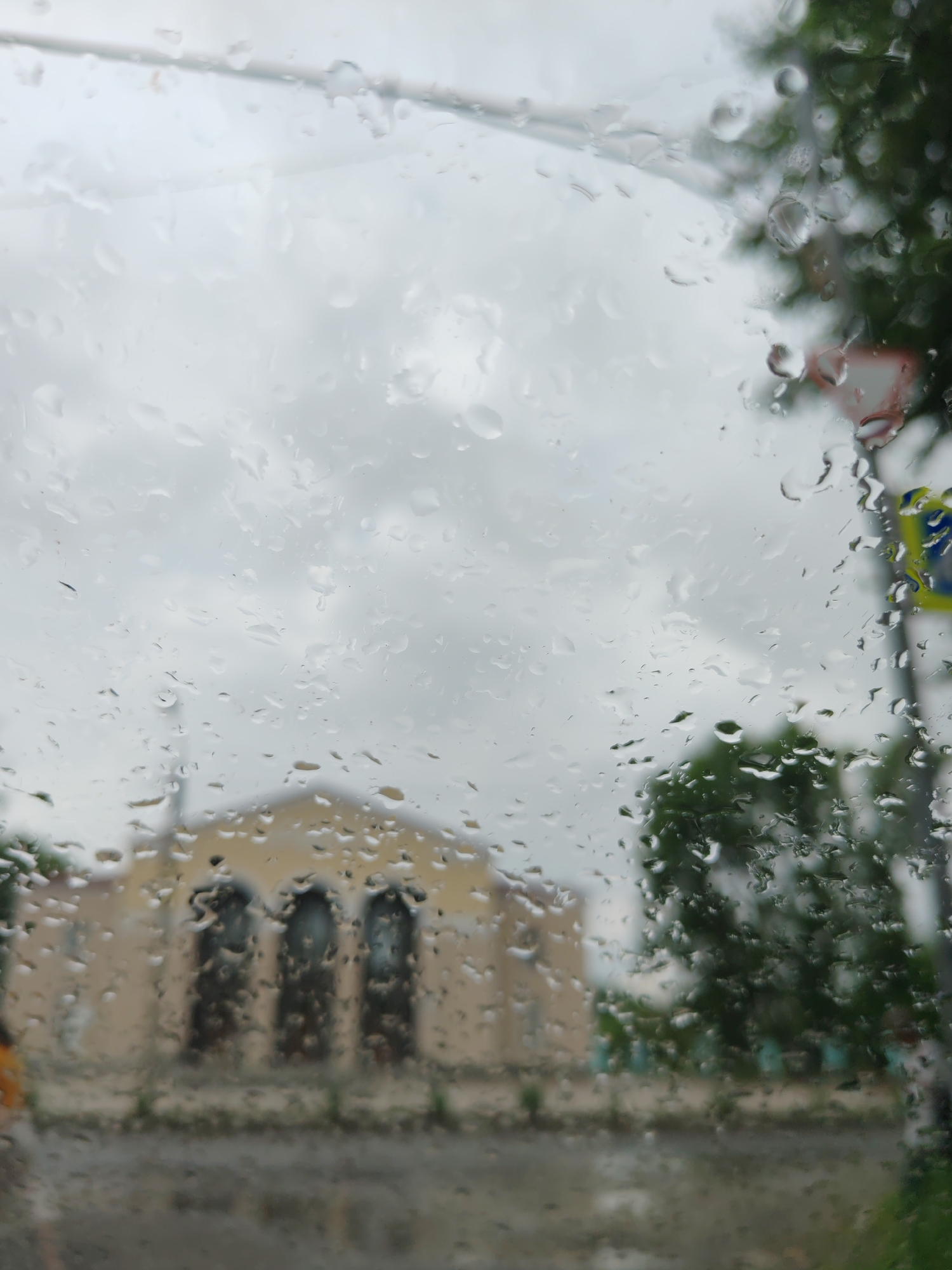 The image depicts a view through a rain-streaked window of what appears to be a building with arched windows, possibly a church or public building, located in Komsomolsk-on-Amur, Russia. The building is light yellow with dark arched openings. In the background, green trees are visible, along with a cloudy, overcast sky. The foreground is dominated by numerous water droplets on the glass, obscuring some of the details of the scene outside. A traffic sign, partially visible and blurred, suggests the location is along a street. No people or specific actions are discernible due to the weather conditions and the quality of the image.