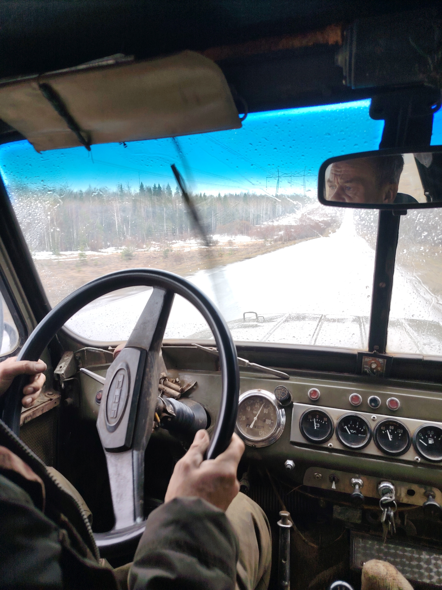 The image depicts the interior of a vehicle, likely a vintage off-road vehicle, from the driver's perspective. A person is shown holding the steering wheel, while the rearview mirror reflects their face. The dashboard features several gauges and switches. Outside the windshield, the scene shows a snow-covered road or path leading through a forest. The presence of utility poles in the background may suggest a rural or semi-rural area. The location is specified as Chaikovsky, Russia, suggesting this is the setting for the scene.