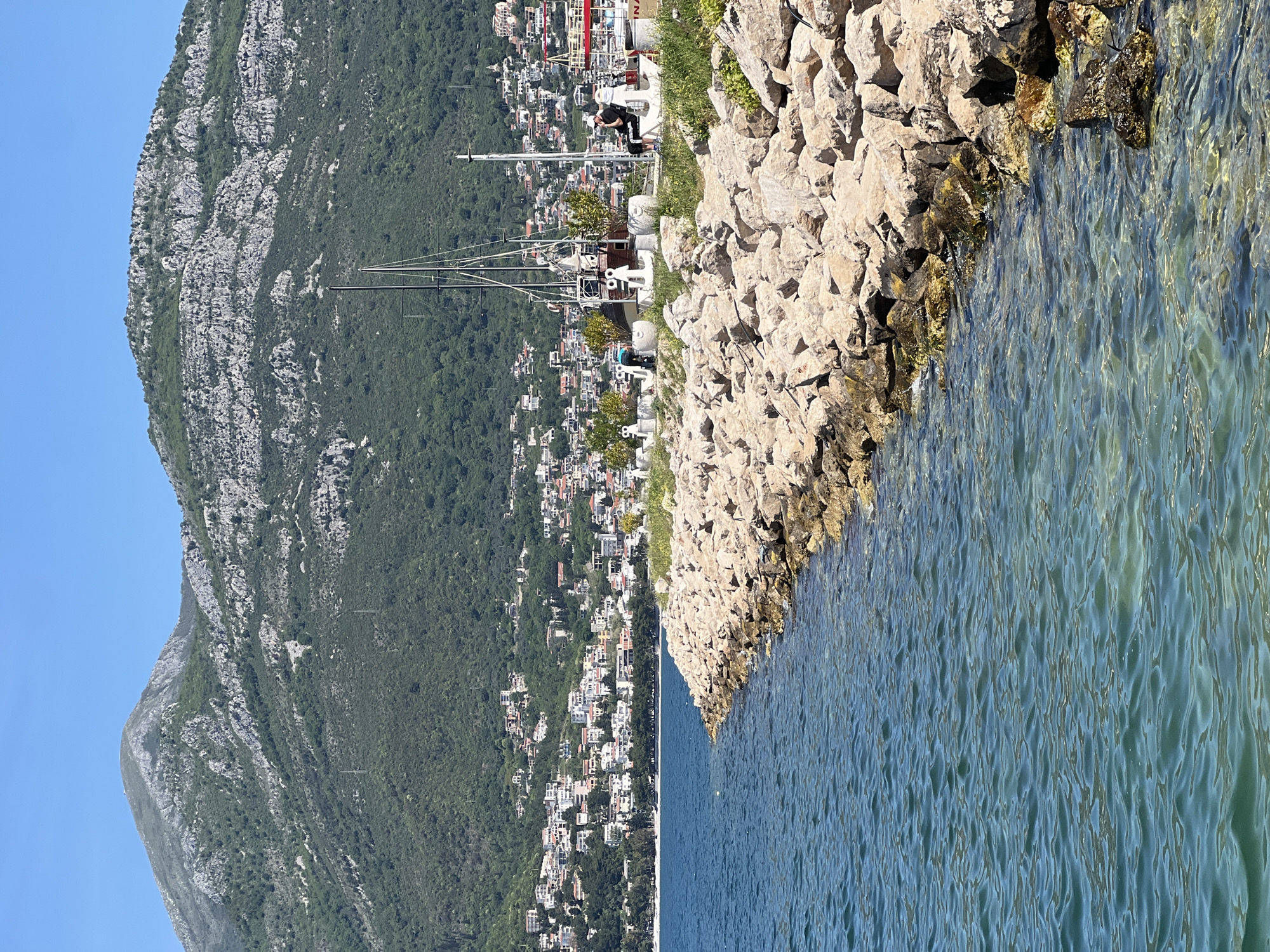 The image shows a rocky shoreline in Bar, Montenegro, bordered by the Adriatic Sea on the right. A long, man-made rock barrier stretches along the coast. Behind the rocks, the city of Bar rises on a hillside covered in lush vegetation. Several buildings are visible, mostly white, intermixed with green trees. On the coast, there are several yachts with tall masts. A mountain rises dramatically in the background. A man is standing, or kneeling on the rocks near a boat. The water is clear, with ripples and some seaweed near the rocks. The sky is blue and clear.