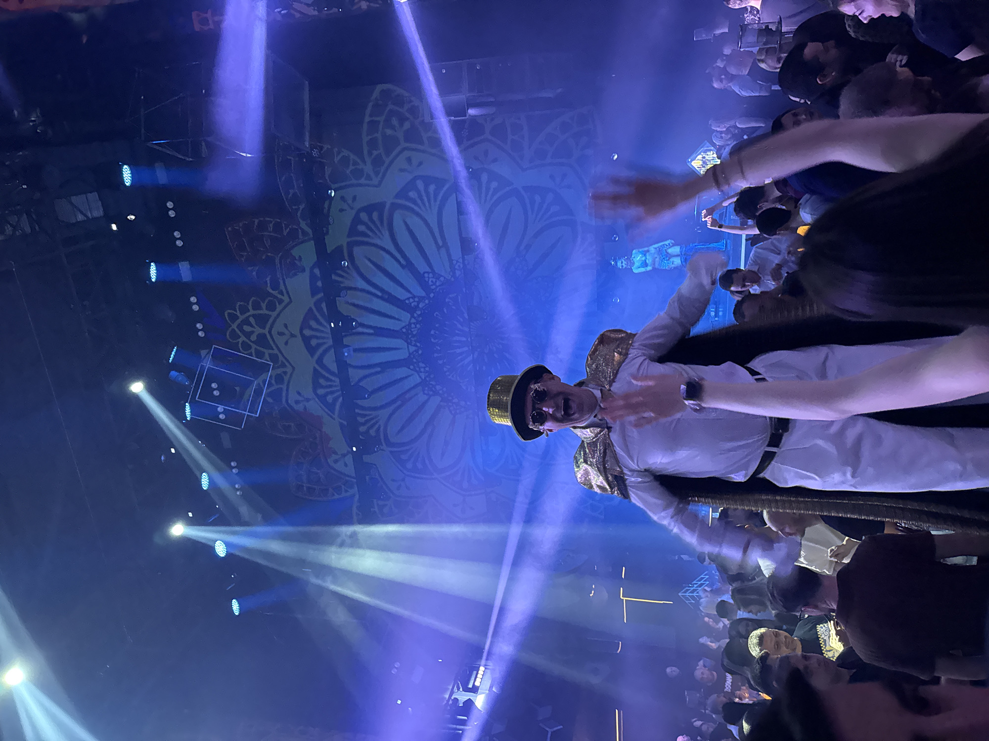 A man in a white suit, gold top hat, and gold cape is standing on a stage in what appears to be a club or concert venue. He is surrounded by a crowd of people. The stage is lit with blue spotlights. A large circular design is visible behind the stage. The location is likely a club or concert venue in the city of Sofia, Bulgaria.