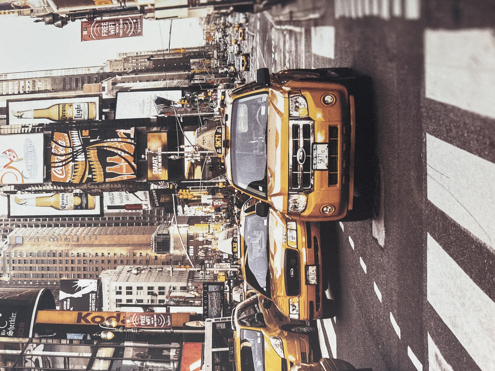 This is a photograph of a busy city street scene, likely in New York City given the presence of yellow taxis and prominent billboards. The image captures a close-up perspective of several yellow taxis, with one prominently featured in the foreground. The taxi in the foreground is orange-yellow and has a license plate that reads "5K71c". The vehicle behind it is also yellow and its license plate is partially obscured but appears to read "6L32A". In the background, towering buildings with numerous windows and advertisements are visible, creating a sense of urban density. Traffic lights and street signs are also present, indicating an active intersection. The ground is paved with asphalt and a white pedestrian crossing is clearly marked. There are no people visible in the immediate foreground, but the overall scene suggests a bustling urban environment with vehicular traffic and pedestrian activity. The image does not depict the city of Anckudinivka, Russia.