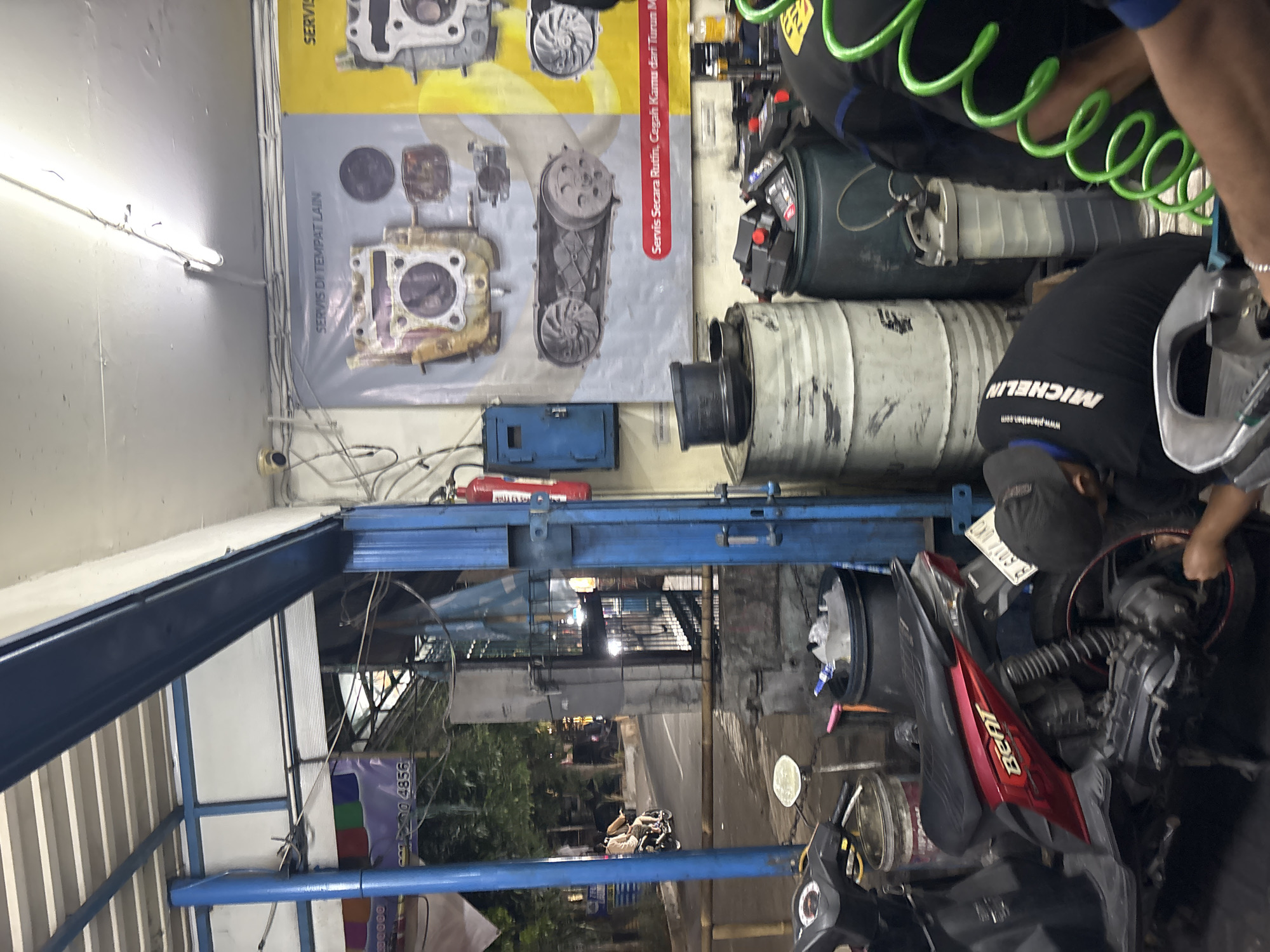 The image depicts the interior of a motorcycle repair shop in North Jakarta, Indonesia, likely on a street with moderate traffic. In the foreground, a red and black Honda Beat motorcycle is parked. A mechanic wearing a black shirt with "MICHELIN" prominently displayed on the back is working on the rear wheel of the motorcycle. To the right, another mechanic is visible, and a bright green coiled hose is prominent in the frame.
The shop environment is functional and appears somewhat cluttered. Large industrial barrels are stacked in the background, one appearing to be a metal drum and another a dark green plastic barrel. Various car parts, possibly engine components, are displayed on posters on the wall, with text in Indonesian that reads "SERVIS DI TEMPAT LAIN" (Service Elsewhere) and "Servis Secara Rutin, Cegah Kerusakan dari Turun Mesin" (Routine Service, Prevent Engine Damage).
A blue electrical cabinet is mounted on the wall, and a red fire extinguisher is also visible. In the mid-ground, through an open doorway, a street scene is visible with trees, buildings, and a couple riding a motorcycle on a road. The lighting suggests it might be late afternoon or evening, with artificial lights illuminating the interior of the shop. Several other motorcycles and miscellaneous items associated with a repair shop are present but less distinct.