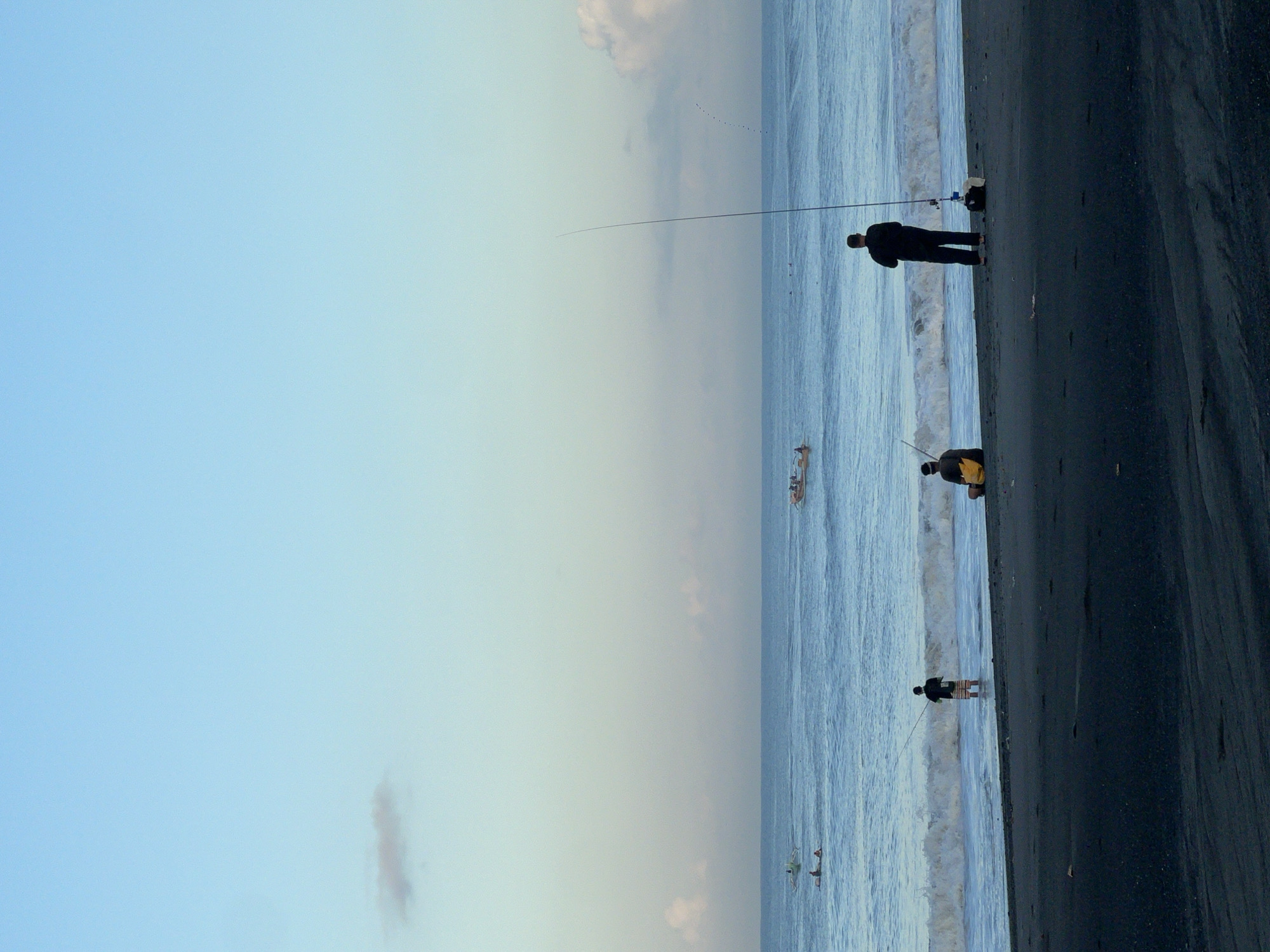 The image depicts a coastal scene in Unknown, Indonesia, showing three figures fishing from the beach. The beach is a dark color, possibly black sand. The sea is a light blue with visible waves. The sky is a gradient from light blue to a pale yellow-gray. In the distance, a ship is visible on the water. The fishermen are silhouetted against the sea and sky. One fisherman is wearing dark clothing, and another is wearing a yellow shirt. All three figures are positioned along the water's edge.