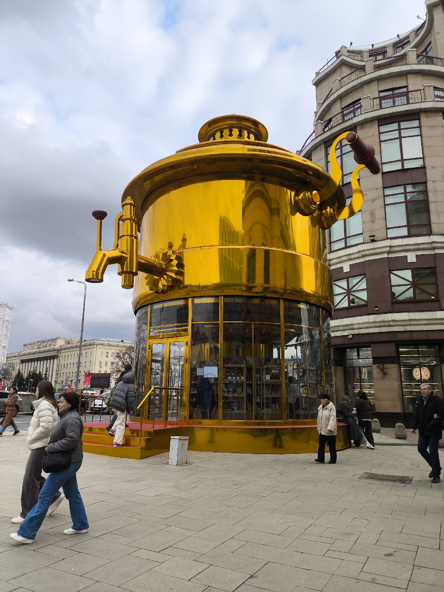 This urban photograph captures a unique, oversized golden samovar-shaped building in Moscow, Russia, dominating the foreground of a busy street scene. The structure is highly reflective, appearing as a giant, polished golden tea urn with a large, decorative spigot/tap on its left side and an ornate handle on its right. The lower part of the samovar forms a circular storefront with glass windows, through which shelves stocked with merchandise are visible. A small set of golden stairs leads up to the shop's entrance.
The weather is overcast, with a cloudy sky providing diffused daylight. Several pedestrians are seen walking on the grey-paved sidewalk around the golden building. In the left foreground, two women walk from left to right; one wears a white puffer jacket and dark pants, the other a grey puffer jacket and blue jeans. Near the samovar's entrance, people are seen entering or exiting, including one person in a light jacket ascending the steps. On the right, an older man in a dark jacket and blue jeans walks past.
In the background, an array of city buildings is visible, including a modern, multi-story structure with a curved facade, balconies, and large windows on the right. Further back on the left, a grander, older building with classical columns can be discerned, typical of European city architecture. A streetlight stands tall on the left. The scene depicts everyday activity in a vibrant city environment. No distinct text is clearly legible on any of the structures or signage.