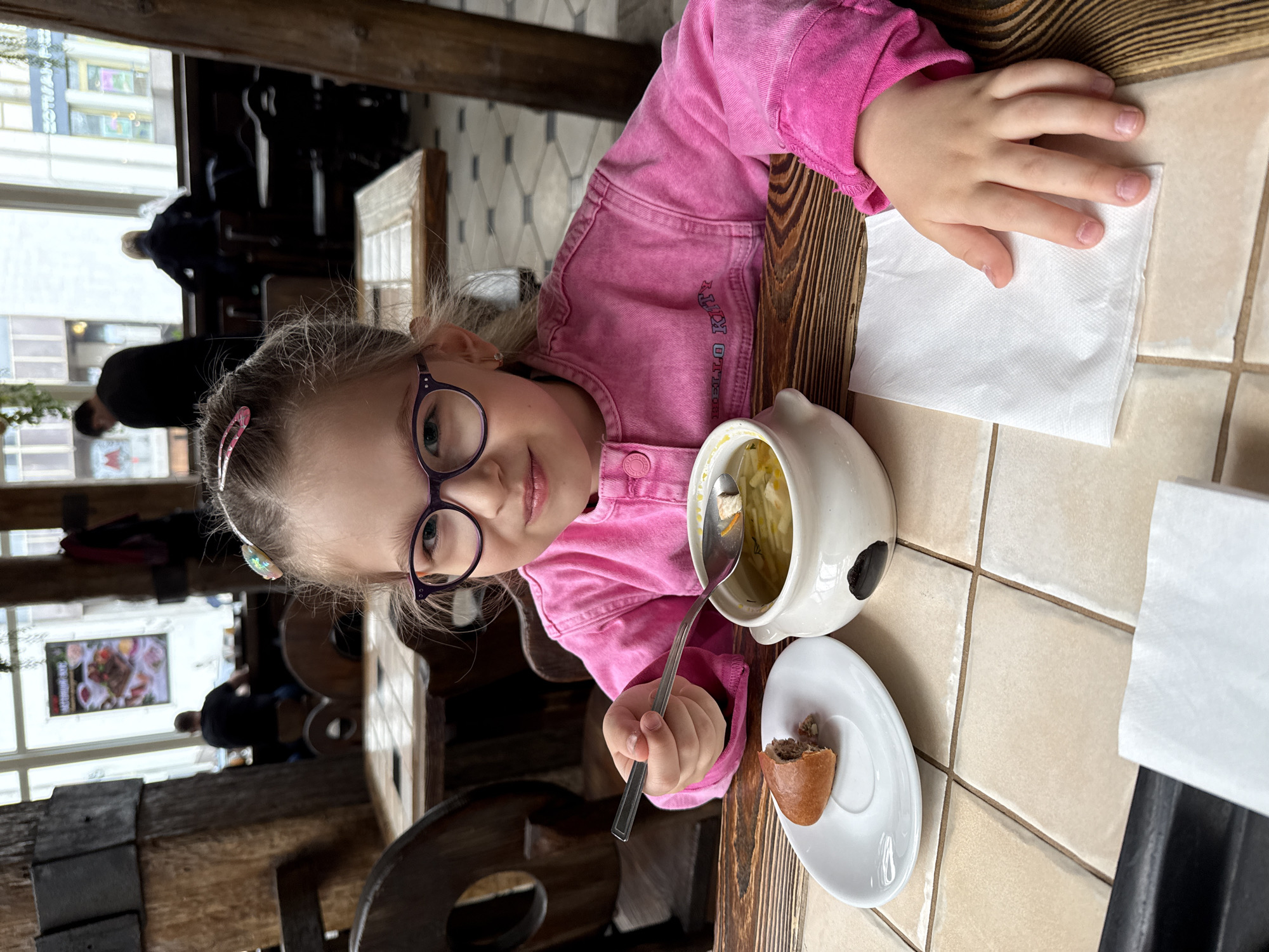 The image shows a young girl with light brown hair, wearing purple-rimmed glasses, a small earring in her right ear, and a pink denim jacket embroidered with "HELLO KITTY" on the front. She is seated at a wooden table with light-colored square tiles, inside a restaurant in Moscow, Russia. The girl holds a silver spoon containing soup, poised above a white ceramic bowl of soup. Adjacent to the bowl, a partially eaten, elongated baked good (pirozhok) rests on a white saucer. A white paper napkin is visible under her right hand. She looks directly at the camera with a subtle smile. The restaurant interior features heavy wooden pillars and dark wooden chairs. In the background, through large windows, a city street is visible, revealing buildings and a blurred sign on an opposing structure that reads "ЗОЛОТАЯ РЫБКА ПИЦЦА" (Golden Fish Pizza) in Cyrillic. Other patrons are indistinctly visible seated at tables in the background. The scene is illuminated by natural daylight from the windows. A poster with Cyrillic text, "НАСТОЯЩИЙ МЯСНОЙ", is visible on a wooden pillar.