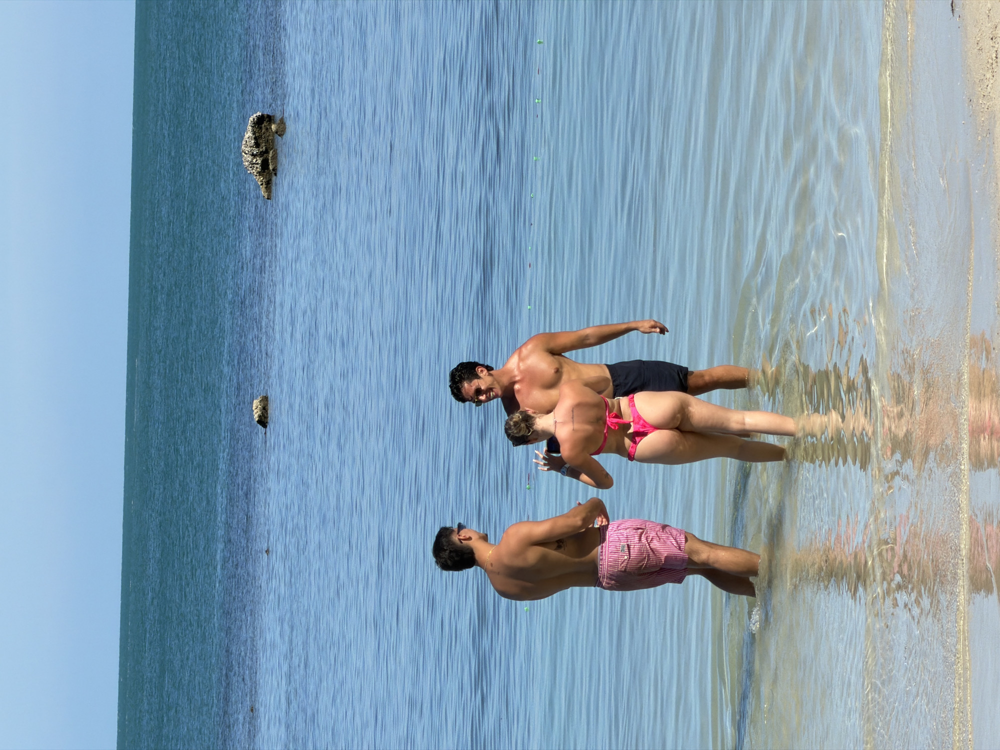 Three people are in shallow, clear blue water near a sandy shore. The sun is bright, suggesting a warm, sunny day, likely midday. The water is calm with gentle ripples and reflections.
In the foreground, a man wearing pink and white striped swim trunks stands facing away from the camera, looking towards the other two people. He has dark hair and is wearing sunglasses.
Further back, a man and a woman are standing closer together. The man, with dark hair, is wearing dark swim trunks. The woman is wearing a bright pink bikini. She is holding a phone in her left hand and appears to be showing something on it to the man. Both are looking down at the phone.
In the distance, a dark, rough object, possibly a rock or piece of coral covered in growth, is visible in the water. Another similar object is visible further out. The background is a vast expanse of blue water and a clear, pale blue sky. The scene evokes a relaxed, leisurely atmosphere typical of a beach vacation. No text is visible in the image.
