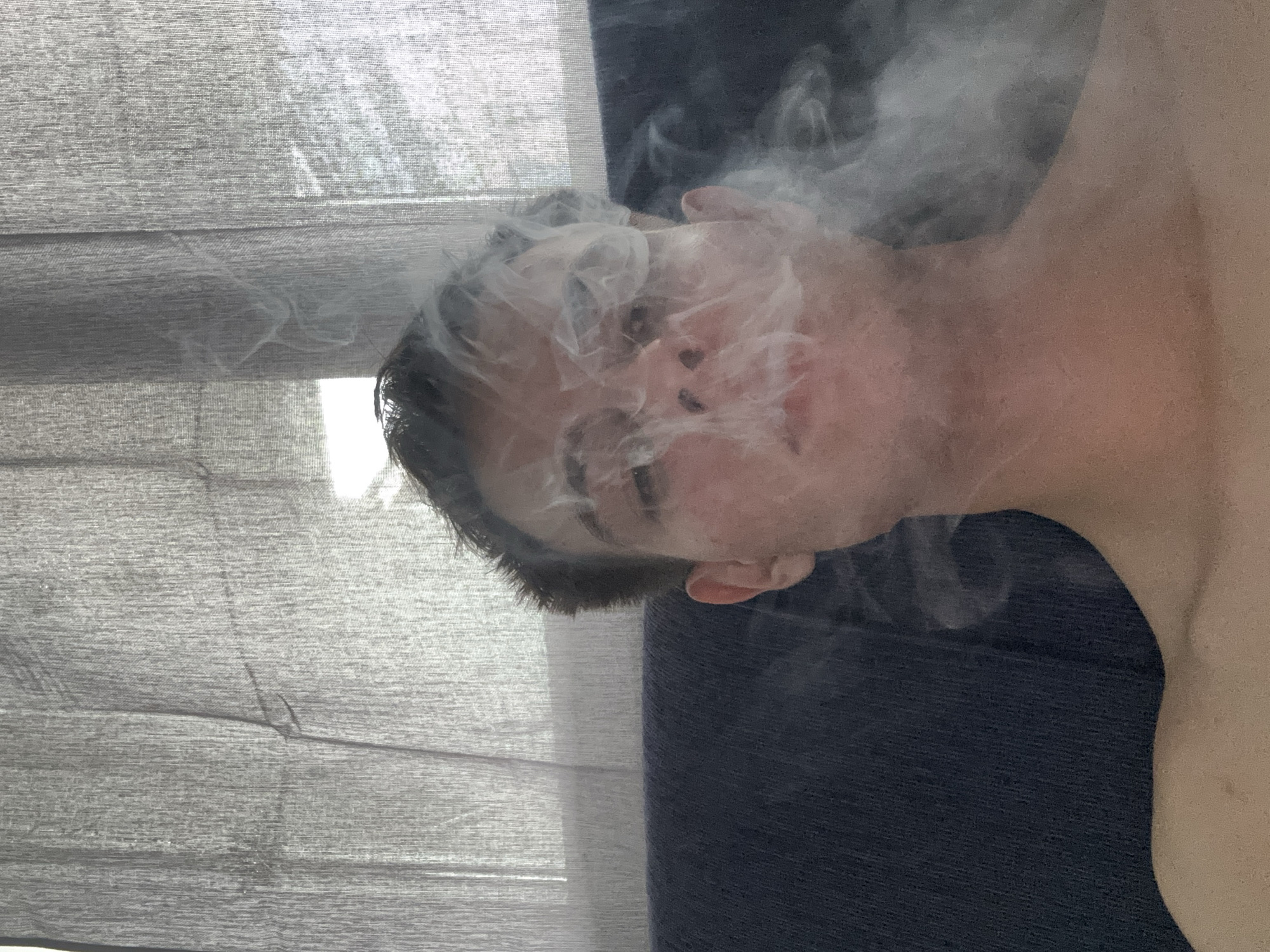 An indoor, portrait-oriented image, rotated 90 degrees clockwise, captures a person's head and upper torso enveloped in a dense cloud of smoke. The individual, with short dark hair, has their face partially obscured by the smoke but appears to be a young adult with a neutral expression. A significant volume of white-grey smoke billows around their head and face, suggesting an act of smoking or vaping. Their bare shoulder and chest are visible on the right side of the frame. The background on the left features light-colored, textured fabric, likely blinds or curtains, through which bright daylight filters. On the right, a darker, solid-colored fabric serves as a backdrop. The scene is set indoors during the daytime. No specific objects or visible text are present, and the overall atmosphere is one of diffused light and hazy visibility due to the smoke.