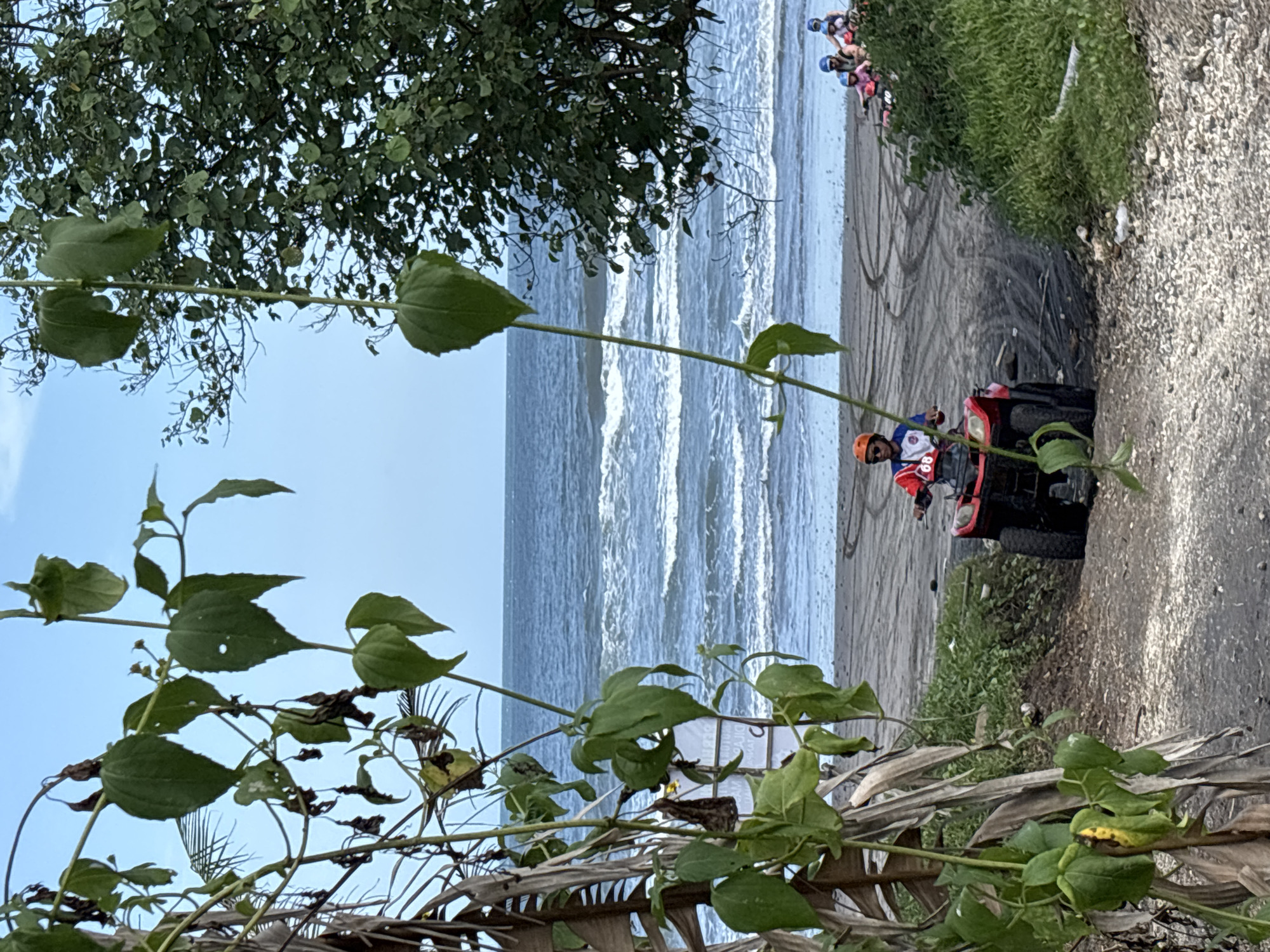 The image shows a beach scene in Kerambitan, Indonesia. In the middle ground, an individual wearing an orange helmet and a racing style jacket is driving a red ATV on the beach. Tire tracks are visible in the sand, indicating recent vehicle activity. Further up the beach, there is a group of people, some wearing helmets, who seem to be either preparing for a ride or observing. The beach itself is adjacent to the ocean, where waves are visible. The background is somewhat obscured by trees and vegetation. In the foreground, there are leafy vines and branches. The overall impression is that of a recreational ATV activity taking place on the beach.