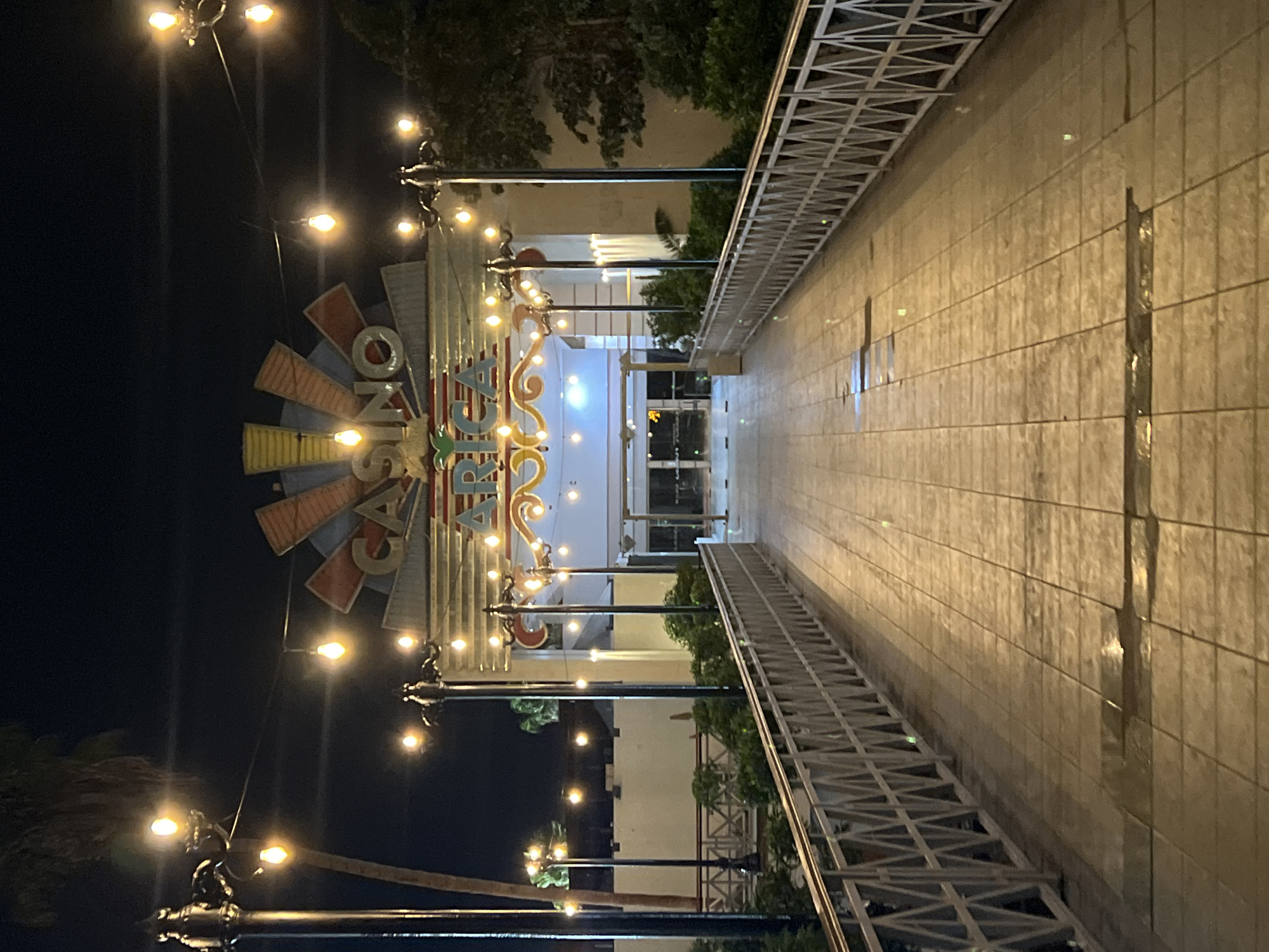 The image depicts the entrance of Casino Arica at night. The casino's illuminated sign is visible, featuring a windmill design. A walkway with metal railings leads to the entrance, which has glass doors. Streetlights line the walkway. The area appears to be outdoors.