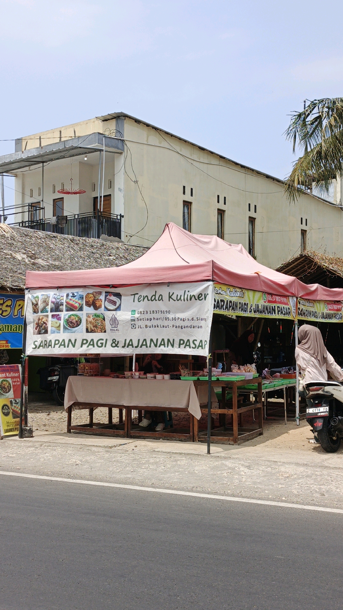 The image shows a street-side food stall, identified as "Tenda Kuliner," located in Pangandaran, Indonesia. The stall is set up under a pink canopy with a banner advertising "SARAPAN PAGI & JAJANAN PASAR" (Breakfast & Market Snacks). The banner also displays images of various food items and contact information, including a phone number (0823 18379190) and an address (JL. Bulak Laut - Pangandaran).
In front of the stall, there are wooden tables set up, covered with beige cloths, displaying food items and containers. Two individuals are partially visible behind the tables, seemingly attending to customers or preparing food. One person is wearing a hijab and a light-colored top, and the other is partially obscured. A motorbike is parked to the right of the stall.
The stall is situated on a paved roadside, with a concrete sidewalk or ground area separating it from the street. Behind the stall, a two-story building with a light-colored facade is visible, featuring several windows and a balcony on the second floor. A portion of a palm tree is also present in the upper right of the frame. The sky is clear and blue.