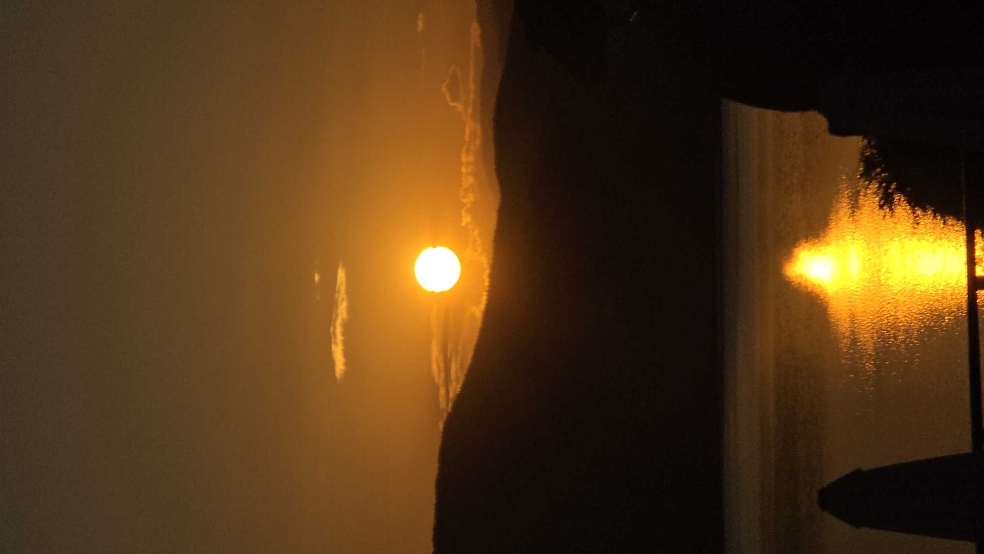 The image depicts a serene sunset over a body of water, with silhouetted hills in the background. The sun, a brilliant orb of light, is positioned centrally in the sky, casting a warm, orange glow across the scene. A small, elongated cloud floats near the sun, its edges illuminated by the sunset. The water's surface is rippled, reflecting the sun's golden light and creating a shimmering path across the scene. In the foreground, the dark silhouettes of what appear to be a beach umbrella or tent and some foliage are visible on the right and left edges of the frame. The overall mood is tranquil and picturesque, capturing the beauty of a natural landscape at dusk. The location of Staro myasto, Bulgaria, is mentioned, but not directly depicted in the image itself.