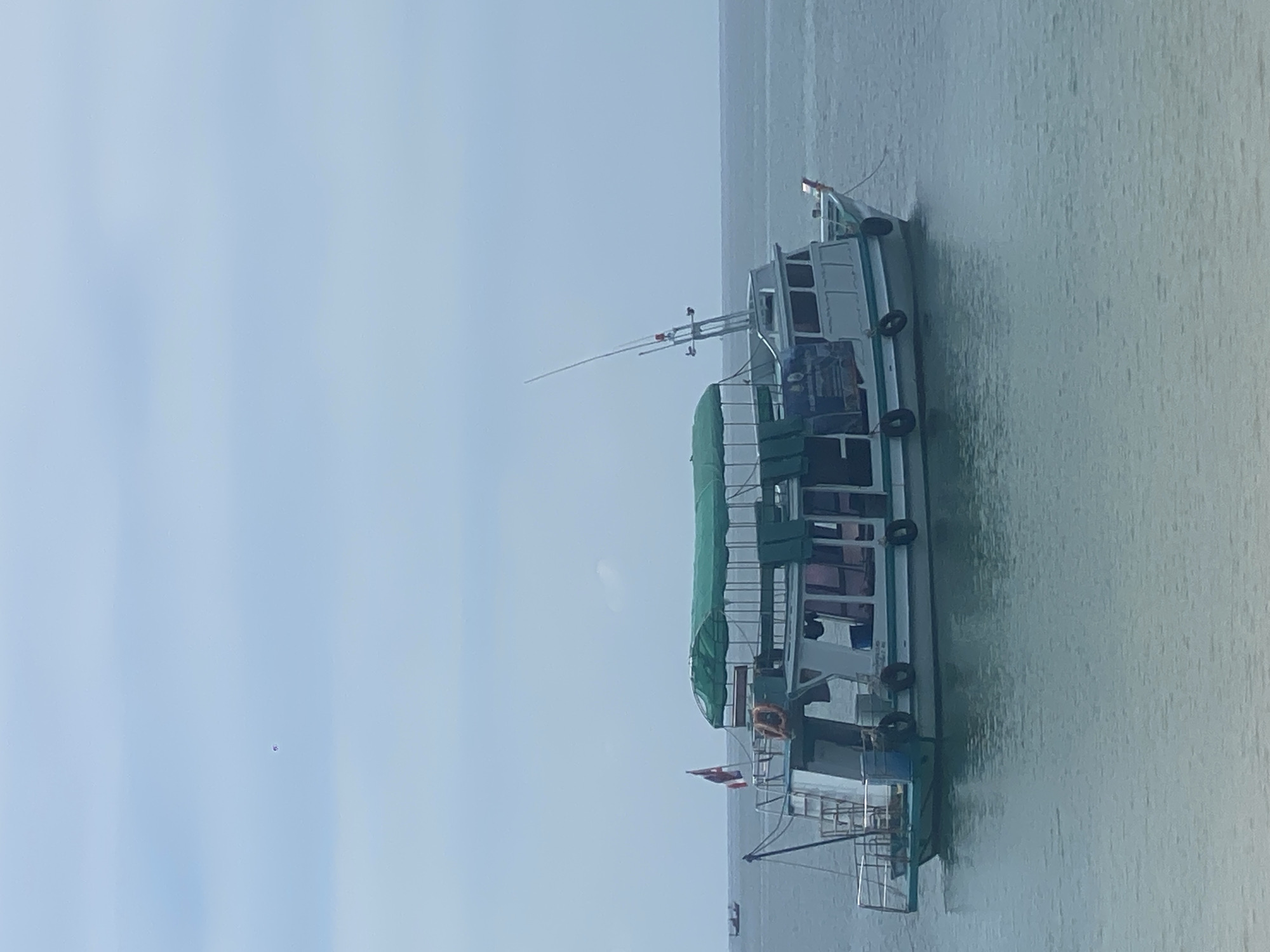 The image shows a passenger boat, presumably in the waters off Baan Chaloklum, Thailand. The boat is white with green accents and has two levels. The upper level is partially covered with a green canopy. There are black inflatable fenders along the side. An antenna extends from the top. A Thai flag is visible at the rear. The sea is calm and the sky is overcast.