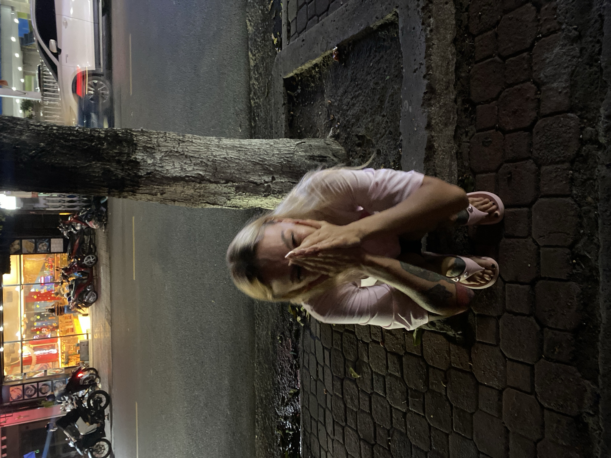 A woman with blonde hair crouches on a brick pavement next to a tree trunk. She wears a light pink shirt and pink sandals. Her hands are covering her face. The pavement is bordered by a road with parked motorbikes and a building with bright lights visible in the background. The scene appears to be at night. The building’s signs suggest the location might be in Đồng Nai, Vietnam.