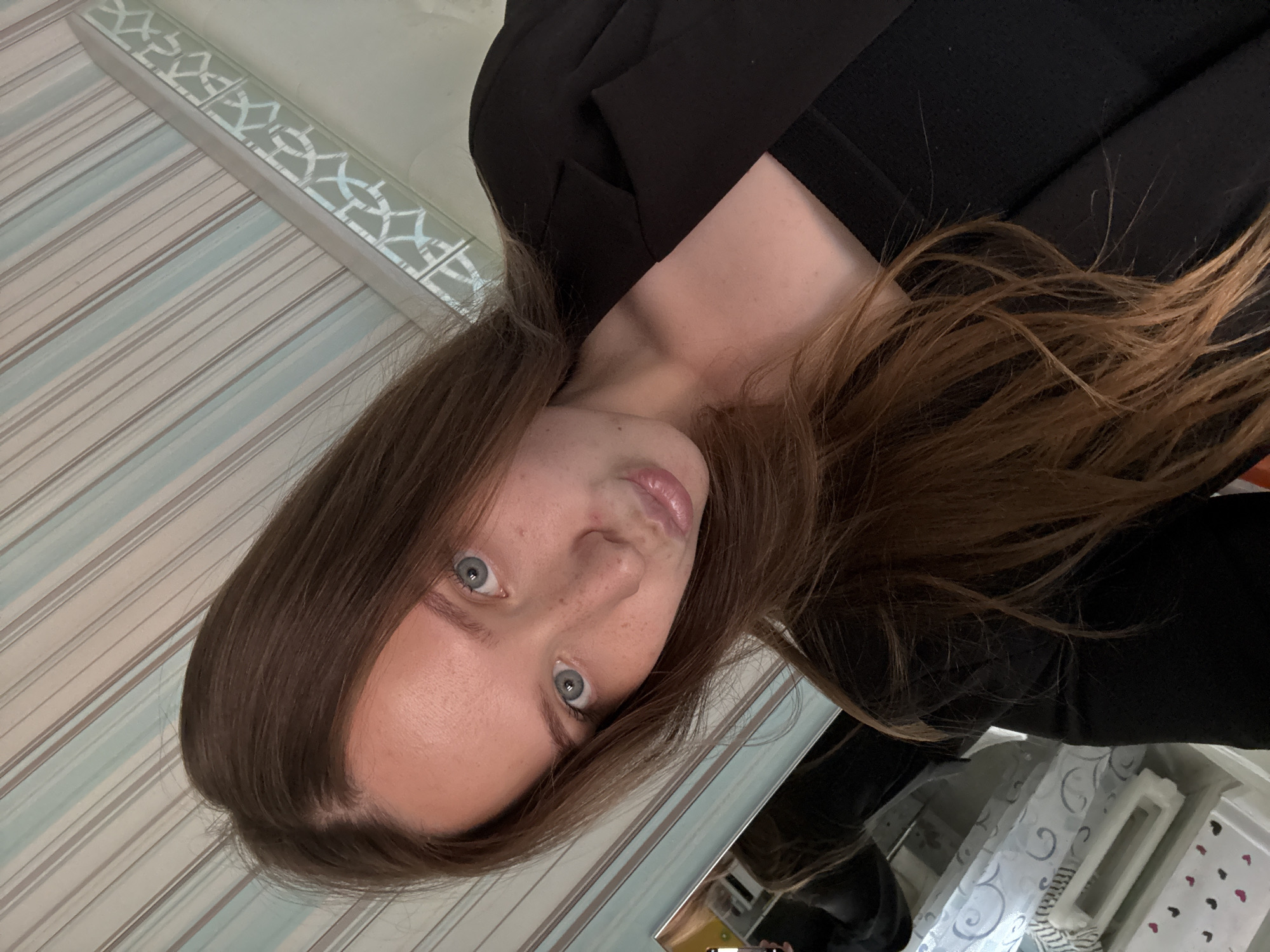 A young woman with long brown hair and blue eyes, wearing a black blazer over a black top, poses for a selfie indoors. She has freckles on her cheeks and nose and her lips are pursed. The background consists of a striped wallpaper and a mirrored surface reflecting part of the room, showing a shelf with decorative items. The location is Chaikovsky, Russia.