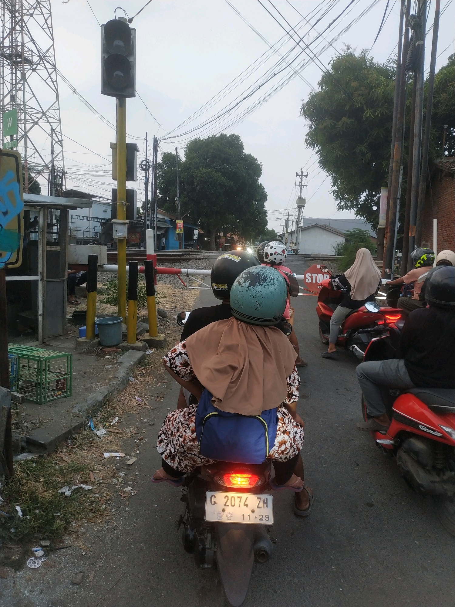 The image captures a scene at a railroad crossing in Tegal, Indonesia. A traffic light, with its red and yellow lenses visible, stands prominently on the left side of the frame. Multiple individuals are waiting on motorcycles, indicating a temporary halt due to the crossing. The foreground is dominated by the back of a motorcycle, displaying a license plate with the characters "G 2074 ZN" and the date "11.29". The riders are wearing helmets, with one person in the foreground wearing a blue helmet and a tan headscarf, and a floral-patterned garment. Other motorcycles with riders are positioned behind, some with visible helmets. The environment suggests a street scene with a railroad track, a barrier, and a power pole with numerous wires. Trees and buildings are visible in the background, contributing to the urban setting.