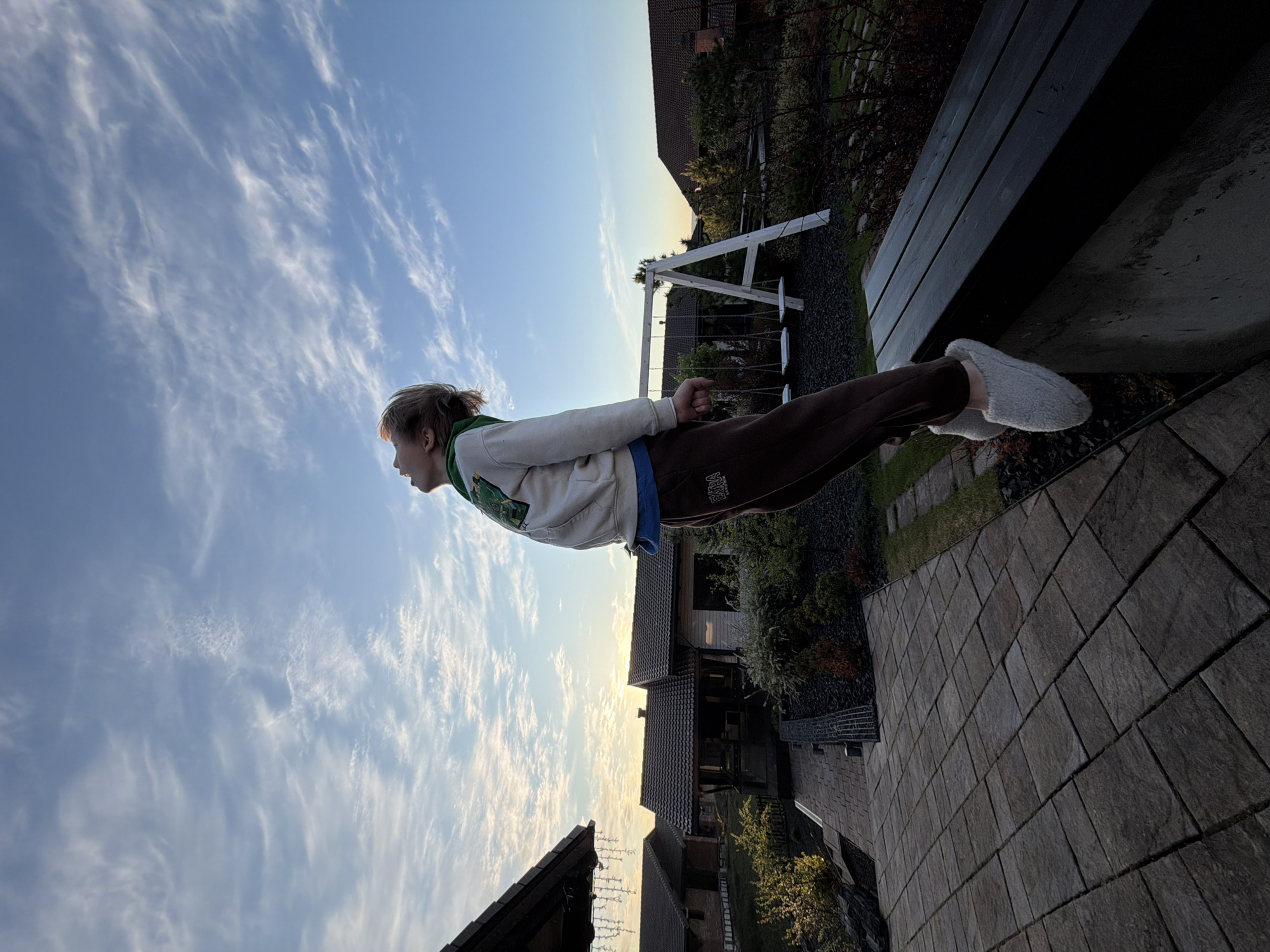 A photograph shows a person lying horizontally on a bench, giving the illusion of flight. The person is wearing brown sweatpants, a light-colored hooded sweatshirt, and slippers. The bench is wooden and appears to be outdoors, with grass and plants visible in the background. In the distance, there is a swing set and houses, suggesting a residential area in Элита, Russia. The sky is blue with wispy clouds, indicating a clear day. The camera angle and orientation create the effect of the person floating in the sky.