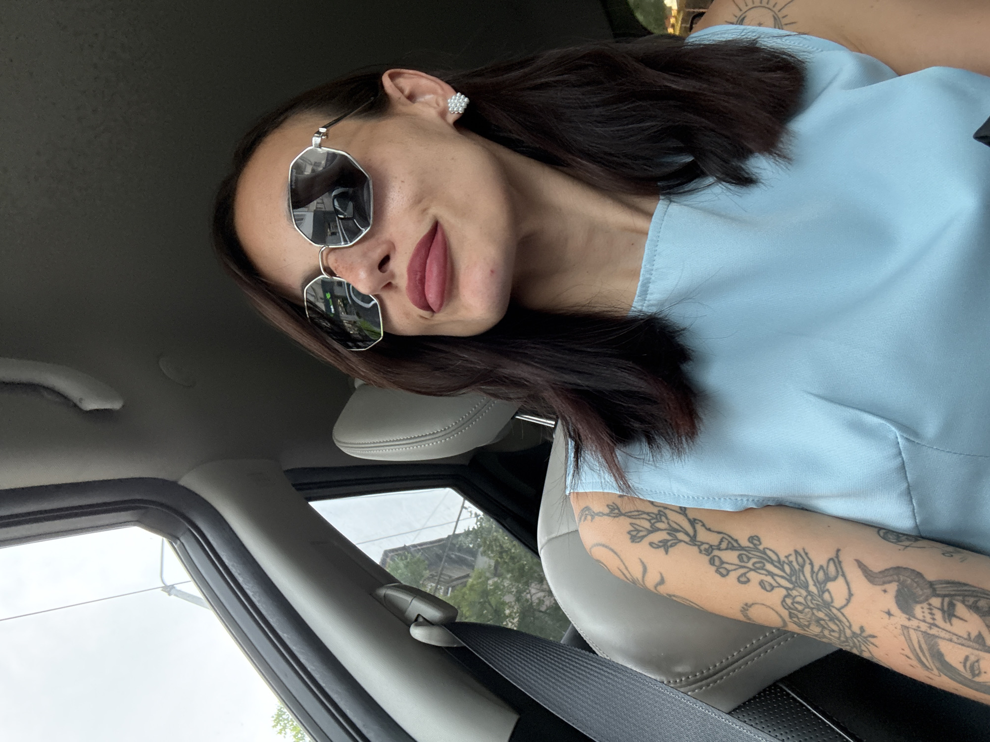The image shows a woman in a car. She is wearing sunglasses and a light blue top. She has tattoos on her left arm and an earring in her left ear. She is wearing dark lip color. The car's interior is visible, including the seat, seatbelt, window, and headrest. Outside the car, buildings and trees are visible through the window. The specific location within the city of Pleven, Bulgaria, cannot be determined from the image alone.