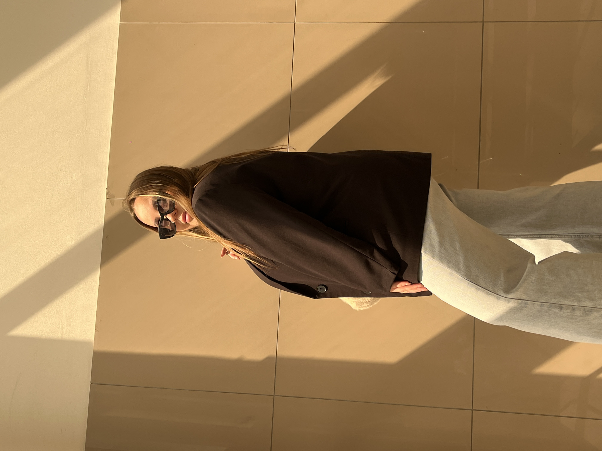 A person with long, light brown hair is wearing sunglasses, a dark blazer, and light blue jeans. The person is standing on a tiled floor in a room with one wall partially visible. The scene is lit by strong sunlight, casting sharp shadows on the floor and wall. The location is Novopokrovskaya, Russia.