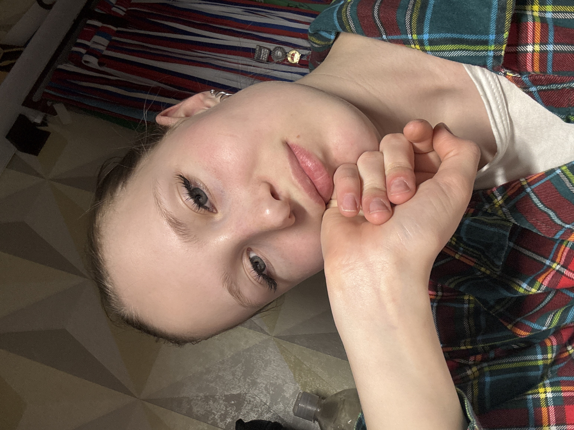 A young, fair-skinned woman with light gray eyes and subtle makeup is lying on what appears to be a geometric patterned floor. She is propped up on her left hand. She is wearing a white undershirt and a plaid shirt with colors of red, green, yellow, white, and blue. A bottle of liquid is near her. In the background, there is a striped textile. The location is Chaikovsky, Russia.
