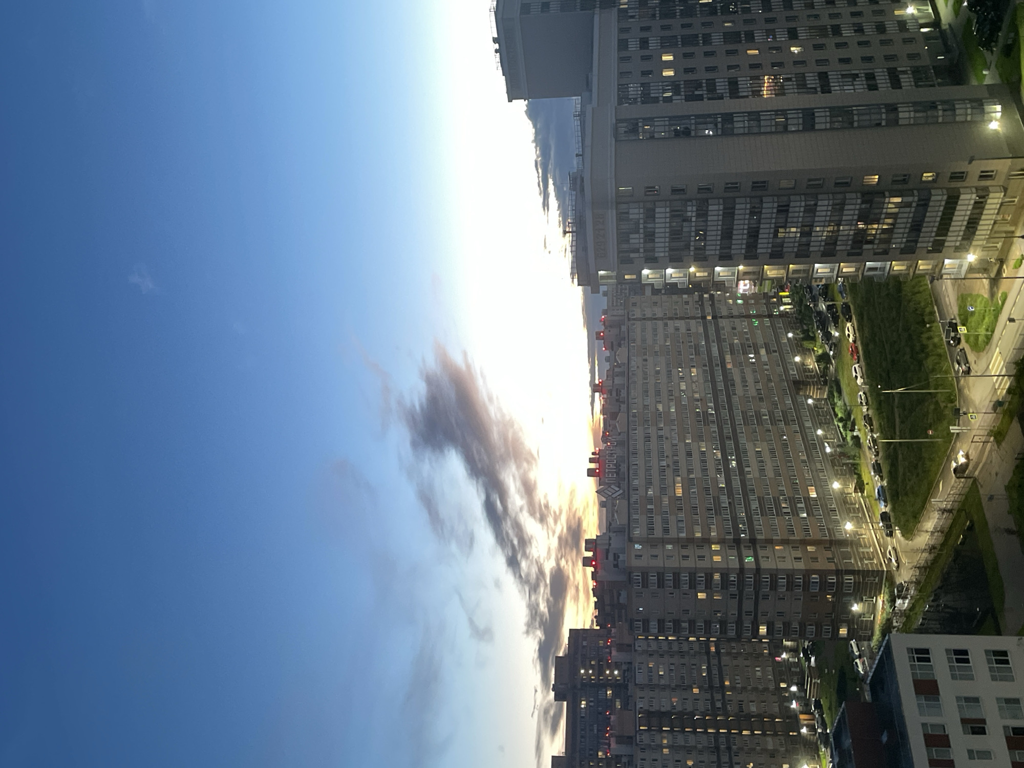 A high-angle, full shot captures a twilight cityscape in an unknown Russian city. The sky is a gradient of deep blue at the top, transitioning to lighter blues and yellows near the horizon, where a few wispy clouds are illuminated by the setting sun. Several large, multi-story apartment buildings dominate the midground and foreground. These buildings are modern, with many windows, some of which are lit, suggesting occupancy. The architecture appears to be of uniform design, common in residential developments. The buildings are closely packed together, creating a dense urban environment. In the foreground, one building stands out with white and red detailing. Streets with parked cars and streetlights are visible at the base of the buildings, indicating urban infrastructure. The overall impression is one of a vast residential area at dusk, with the last light of day illuminating the sky.