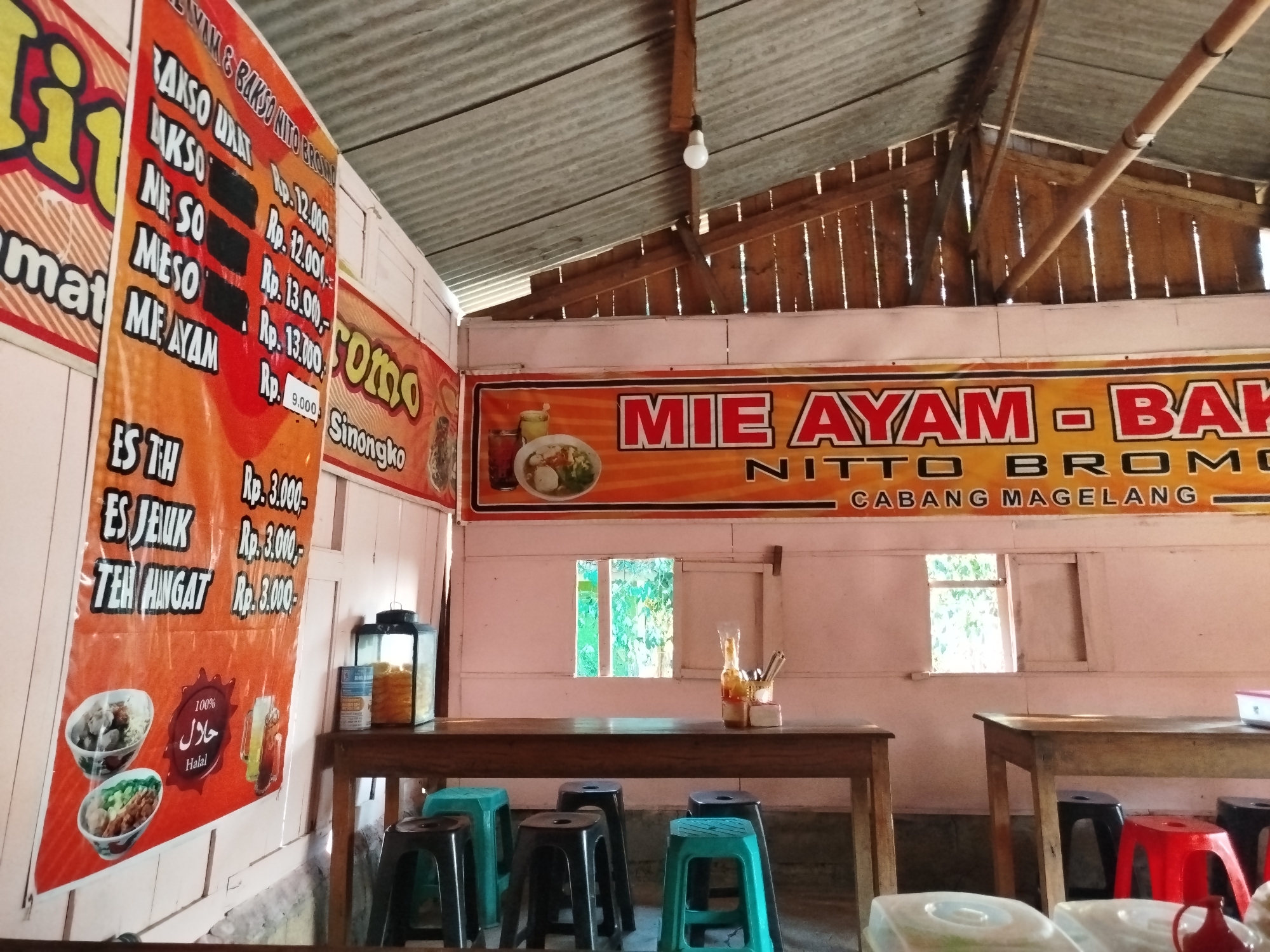 This image depicts the interior of a casual dining establishment, likely a restaurant or food stall, in Pojok, Indonesia. The scene is dominated by a rustic wooden structure with a corrugated metal roof, characteristic of simple buildings in rural or semi-rural Indonesian settings.
In the foreground, a long wooden table is set with several stools of various colors, including black and teal. On the table, there are condiments, including bottles of sauce and a container of what appear to be utensils. To the right, another table is partially visible, also with stools.
Two prominent banners hang on the wall. The banner on the left, predominantly orange, displays a menu with items like "BAKSO URAT", "BAKSO", "MIE SO", "MESO", and "MIE AYAM", along with their prices in Indonesian Rupiah (Rp). It also features images of food and a "Halal" certification. The banner on the right, also orange, announces "MIE AYAM - BAKSO NITTO BROMO CABANG MAGELANG", indicating the name of the establishment and its branch location in Magelang.
The environment suggests a simple, open-air or semi-enclosed dining area. The lighting appears to be a mix of natural light filtering through windows and artificial light from a bulb visible on the ceiling. The overall impression is that of an unpretentious, local eatery serving Indonesian specialties. No people are visible in the image.